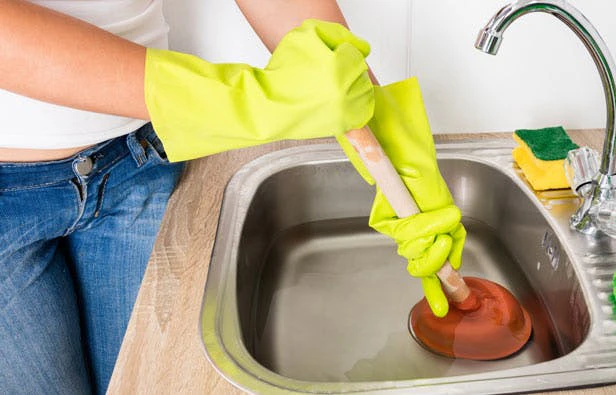 Personne portant des gants en caoutchouc jaunes utilisant une ventouse pour déboucher un évier de cuisine