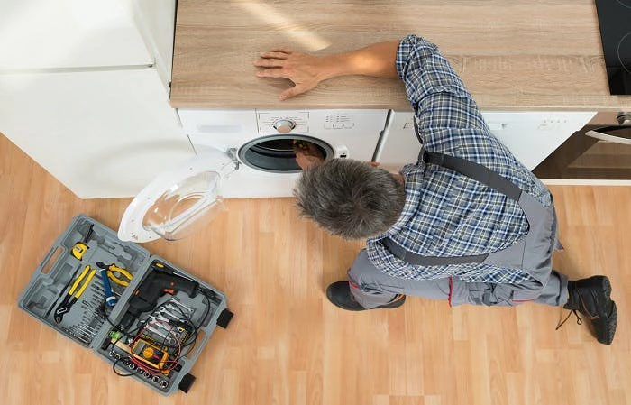 Technicien en salopette réparant un lave-linge avec sa boîte à outils posée au sol