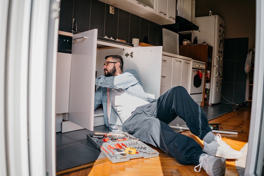 Homme allongé sur le sol de la cuisine inspectant les canalisations sous l'évier avec une boîte à outils