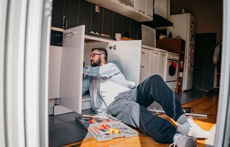 Homme allongé sur le sol de la cuisine inspectant les canalisations sous l'évier avec une boîte à outils
