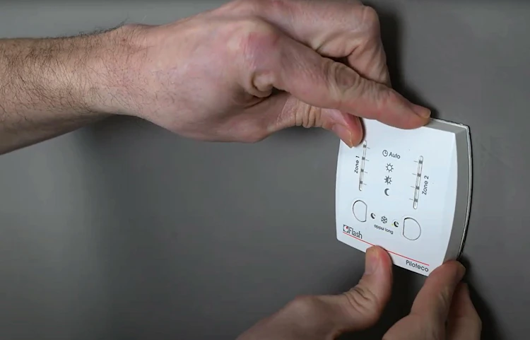 Deux mains fixant un thermostat programmable blanc sur un mur