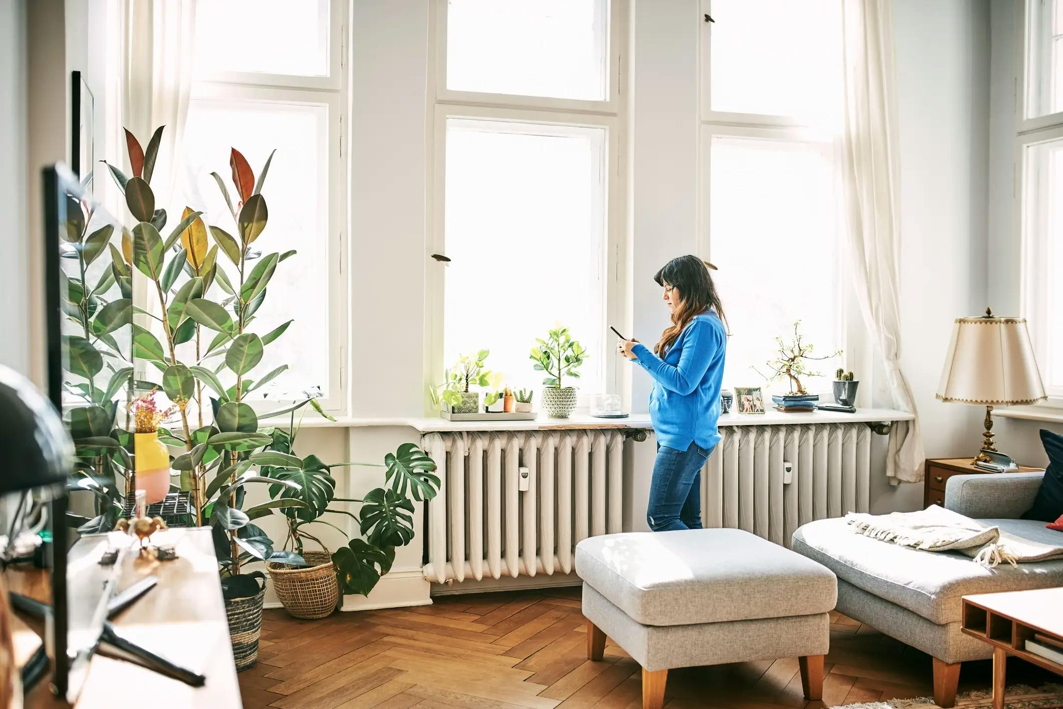 Femme debout dans un salon lumineux, consultant son téléphone devant un radiateur sous les fenêtres