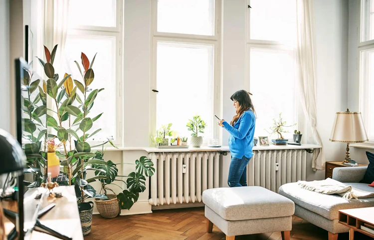 Femme debout dans un salon lumineux, consultant son téléphone devant un radiateur sous les fenêtres