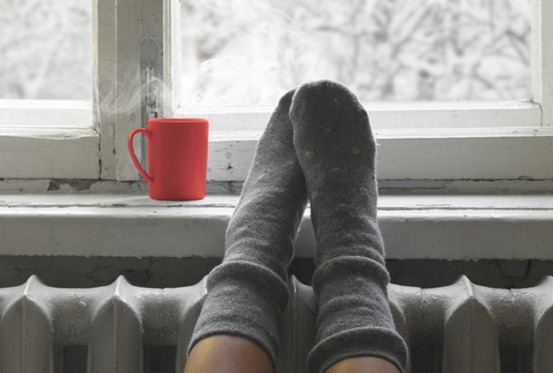Pieds chaussés de chaussettes grises posés sur un radiateur devant fenêtre enneigée