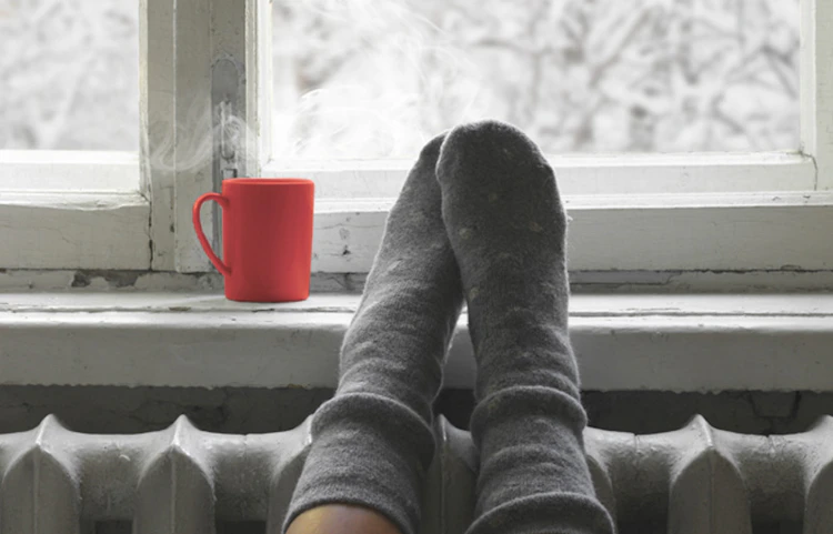 Pieds chaussés de chaussettes grises posés sur un radiateur devant fenêtre enneigée
