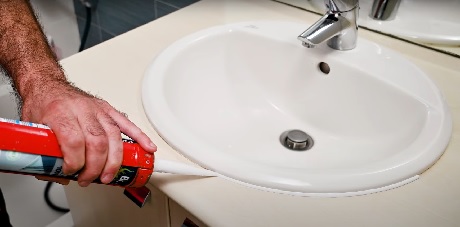 Main appliquant du silicone autour d'un lavabo blanc avec un pistolet à mastic