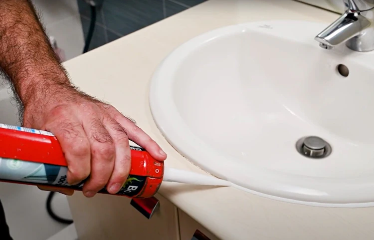 Main appliquant du mastic silicone autour du bord d'un lavabo blanc