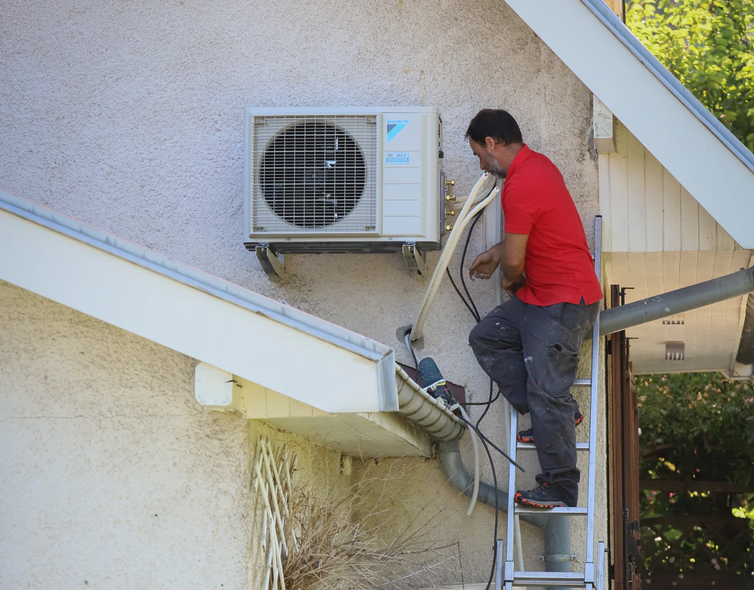 Un technicien VIK Energies installe une unité extérieure de climatisation en façade d'une maison, en hauteur près de La Hague