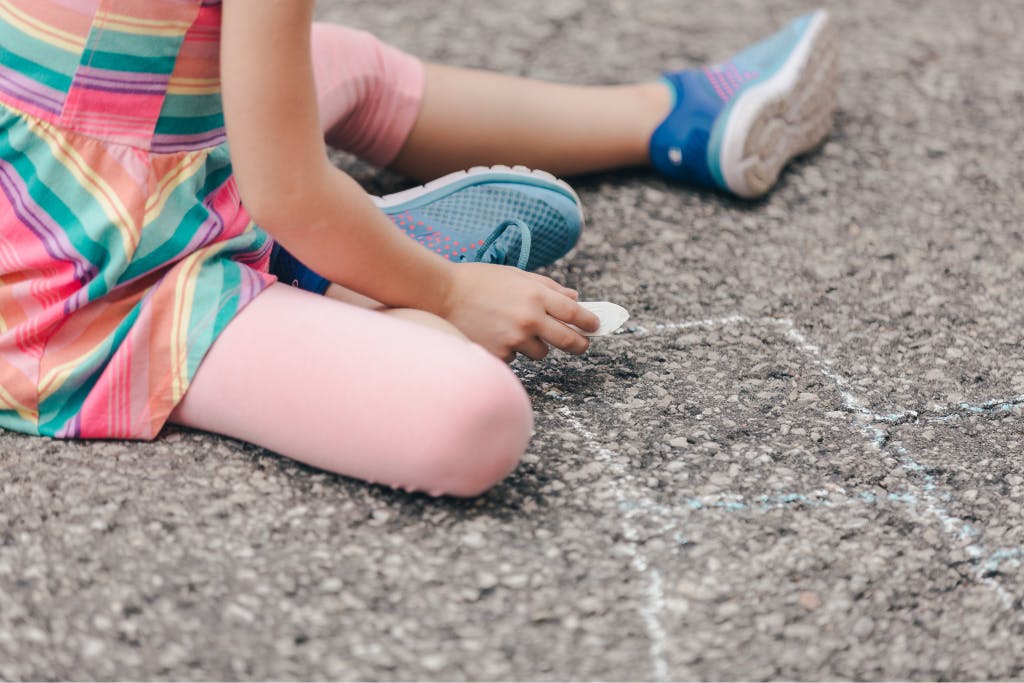 Close-up shot of a child wearing pink tights and blue shoes drawing with chalk on the asphalt.