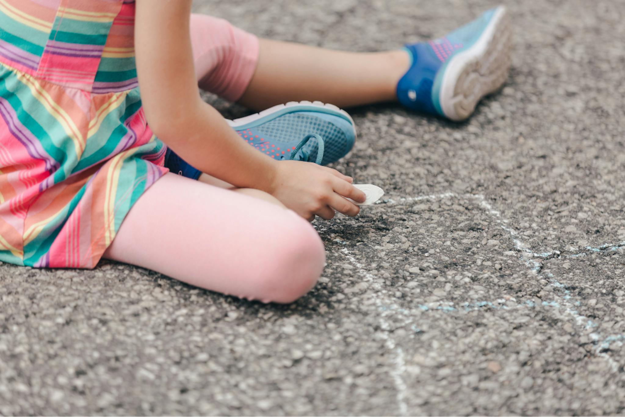 Close-up shot of a child wearing pink tights and blue shoes drawing with chalk on the asphalt.