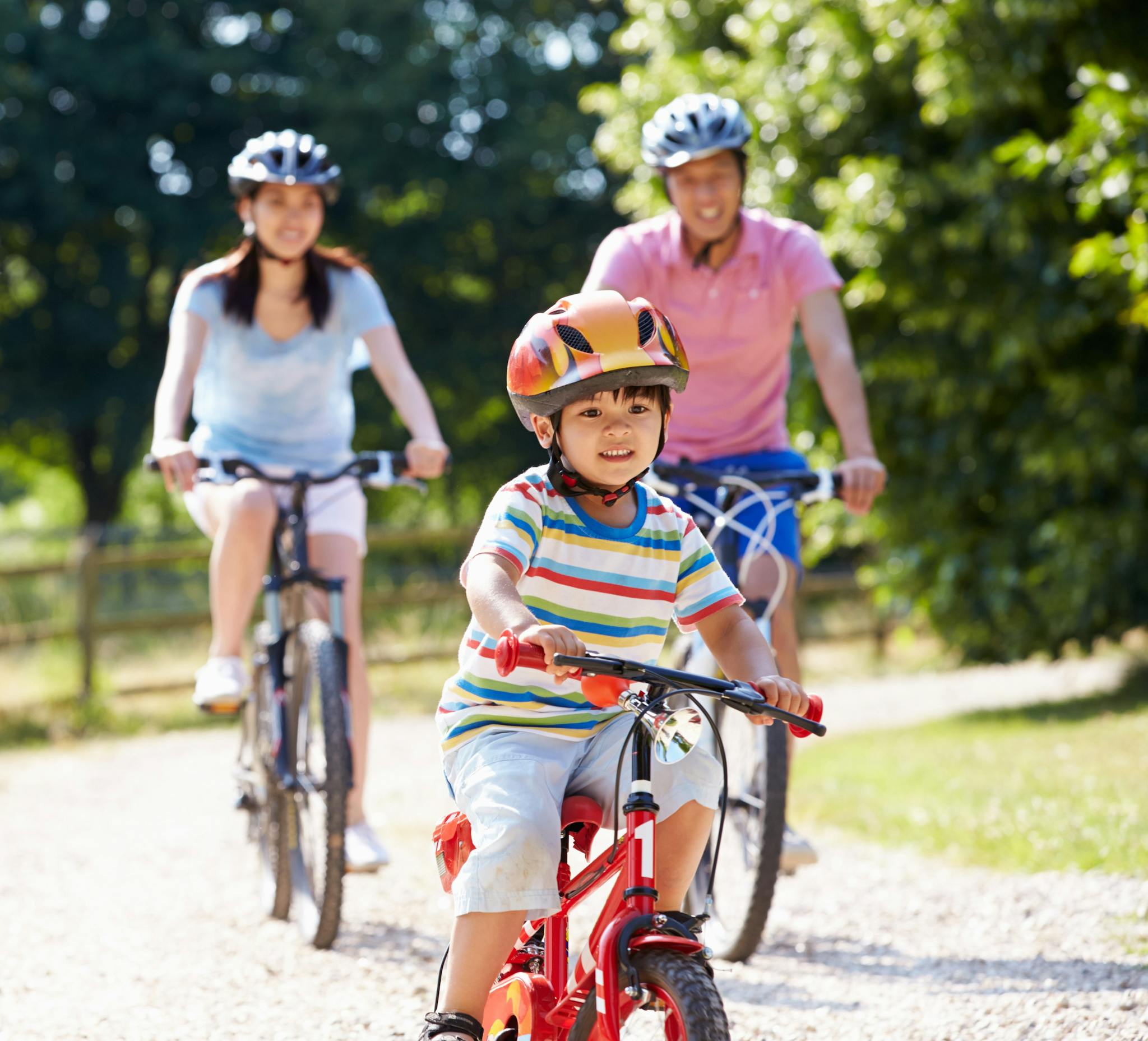 A child riding a red bike down a forested trail, while his parents trail behind.