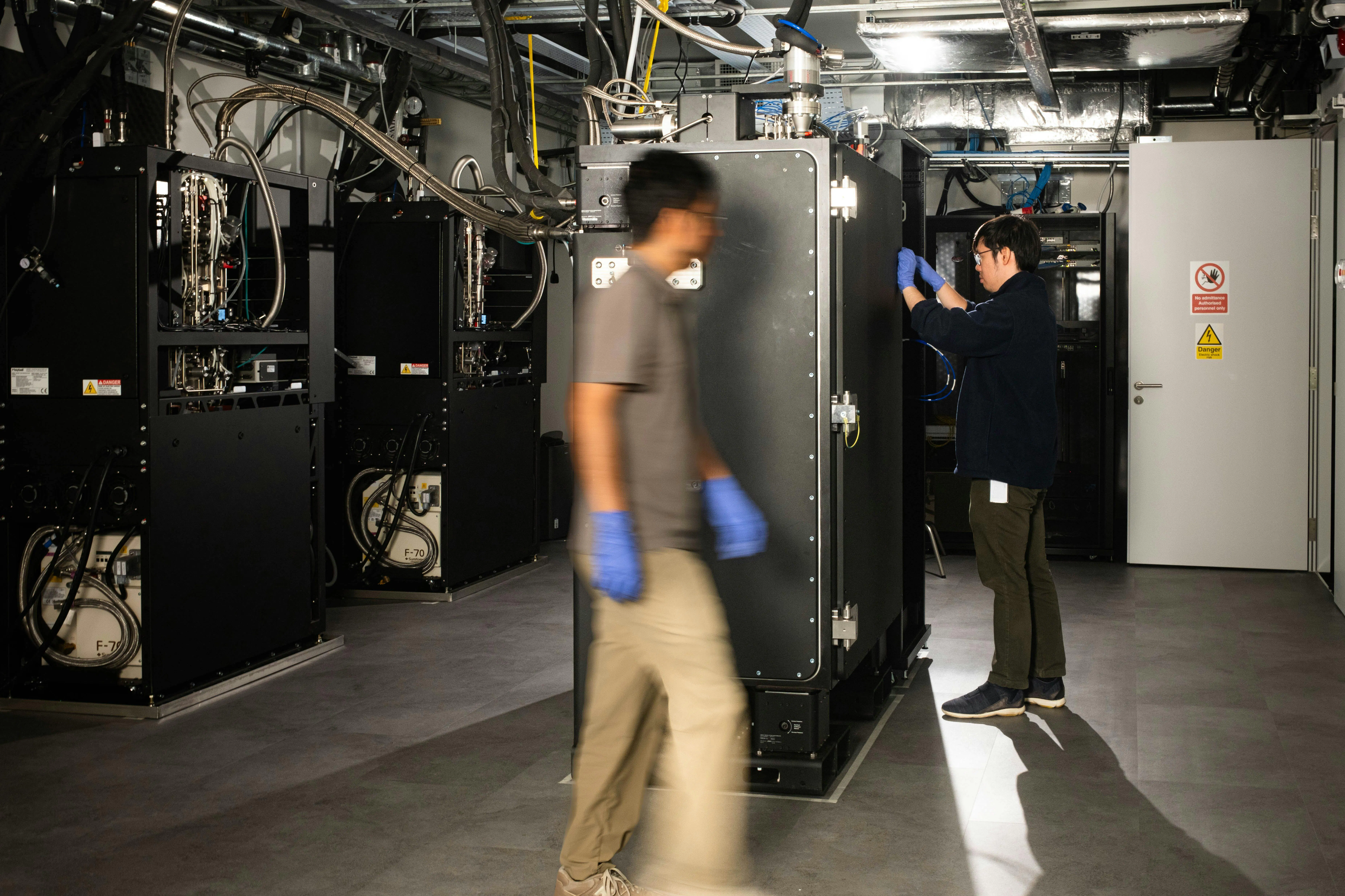 Horizon Quantum staff members in the testbed, assembling the quantum computer, wearing gloves. One walks next to Maybell's Big Fridge, which is closed. The other stands next in front of it as he works on the control system, which is out of view, hidden by the fridge.
