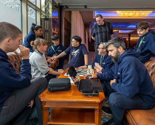 A group of nine Horizon team members relax in a seating area containing two sofas and a table. Some sit on the sofas. Others perk on the arms and back of them. Others stand. On the table in the middle, there is a laptop and one team member is at work on it, with his hand on the mouse. There is also a chess board on the table, set for a game.