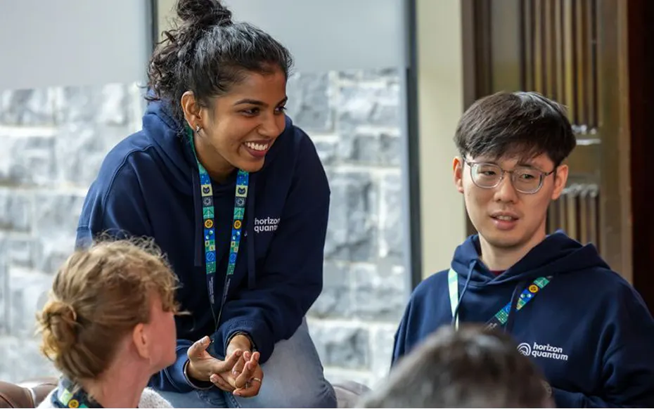 A Horizon team member sits perched on the arm of a sofa. She is laughing. her colleague is standing next to her, and he is smiling.