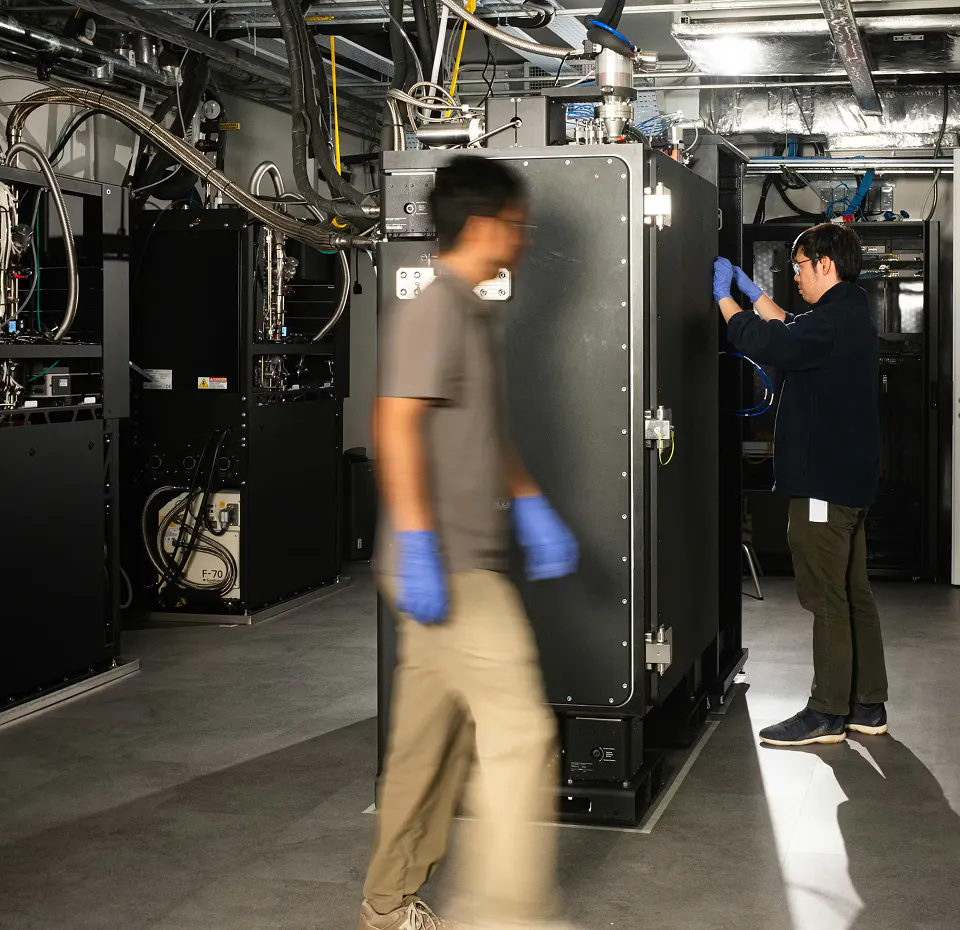 Horizon Quantum staff members in the testbed, assembling the quantum computer, wearing gloves. One walks next to Maybell's Big Fridge, which is closed. The other stands next in front of it as he works on the control system, which is out of view, hidden by the fridge.