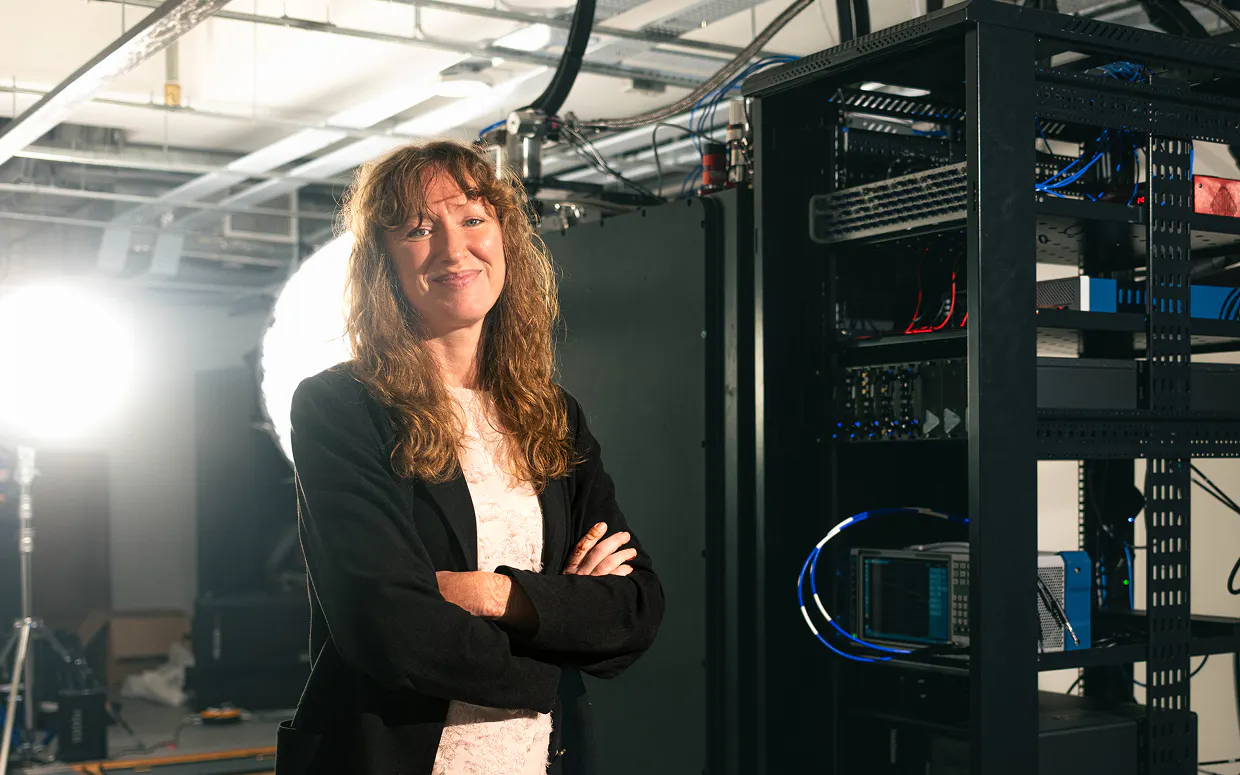 A Horizon team member stands in front of Ember-1, Horizon's quantum computer. She is smiling. There are bright lights behind her. The quantum computer is contained in a large black refrigerating unit.