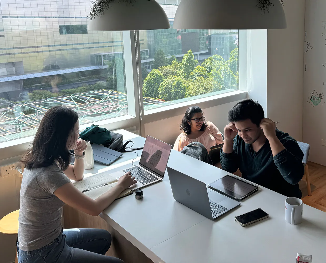 Three Horizon interns working at the counter in the cafeteria. They have laptops, tablets, phones, and coffee cups in front of them. They look relaxed and collaborative. Two are smiling an laughing while the other holds his hands to his ears, pressing EarPods in them with a look of amused focus on his face.