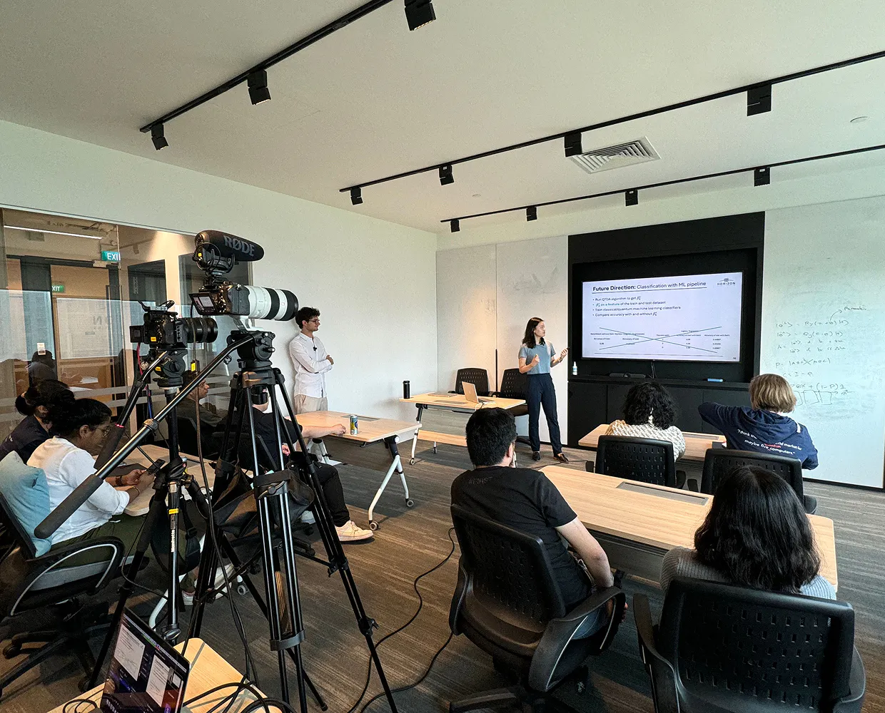 A Horizon intern presents to a room full of people She's standing next to a screen with text on it, which is next to a whiteboard with equations and text on it.
