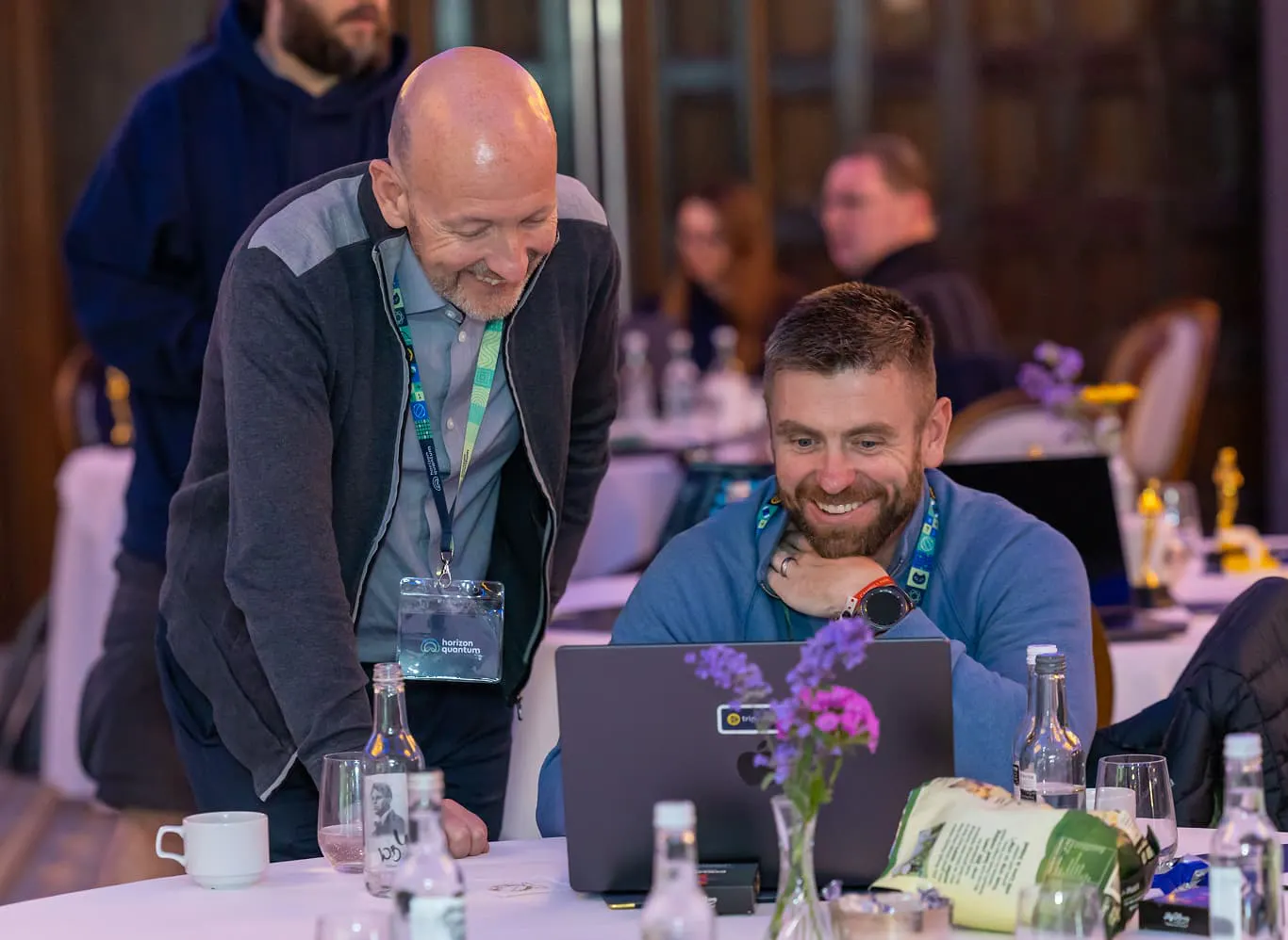 A Horizon team member is staring at his laptop which is on a banquet table. He's laughing while another team member looks over his shoulder, also laughing.