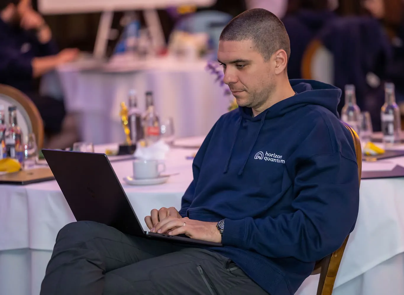 A Horizon Quantum team member sitting next to a banquet table covered in a white tablecloth, typing on his laptop which is resting on his lap.