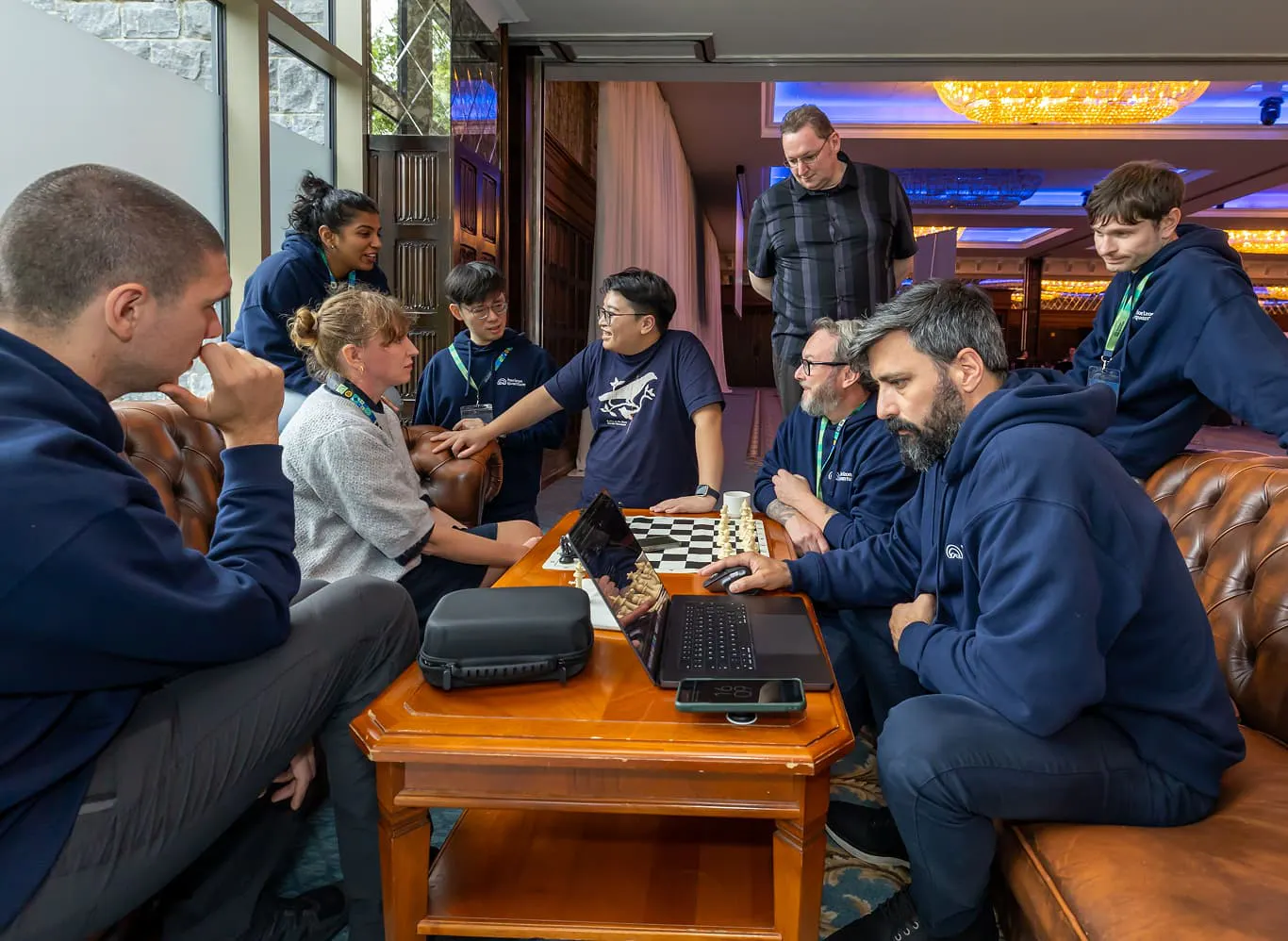 A group of nine Horizon team members relax in a seating area containing two sofas and a table. Some sit on the sofas. Others perk on the arms and back of them. Others stand. On the table in the middle, there is a laptop and one team member is at work on it, with his hand on the mouse. There is also a chess board on the table, set for a game.