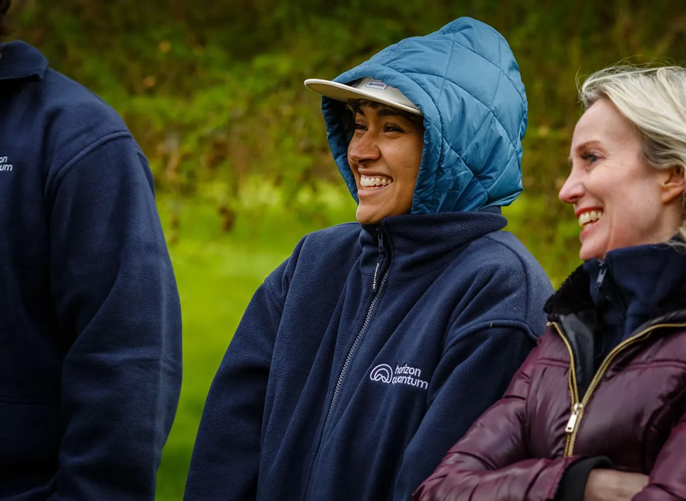 Two Horizon Quantum team members laugh at something we can't see. They're outside at the company offsite. On woman has a hood pulled up over her ball cap, as if it's been raining recently, and the other is bundled up in a coat.