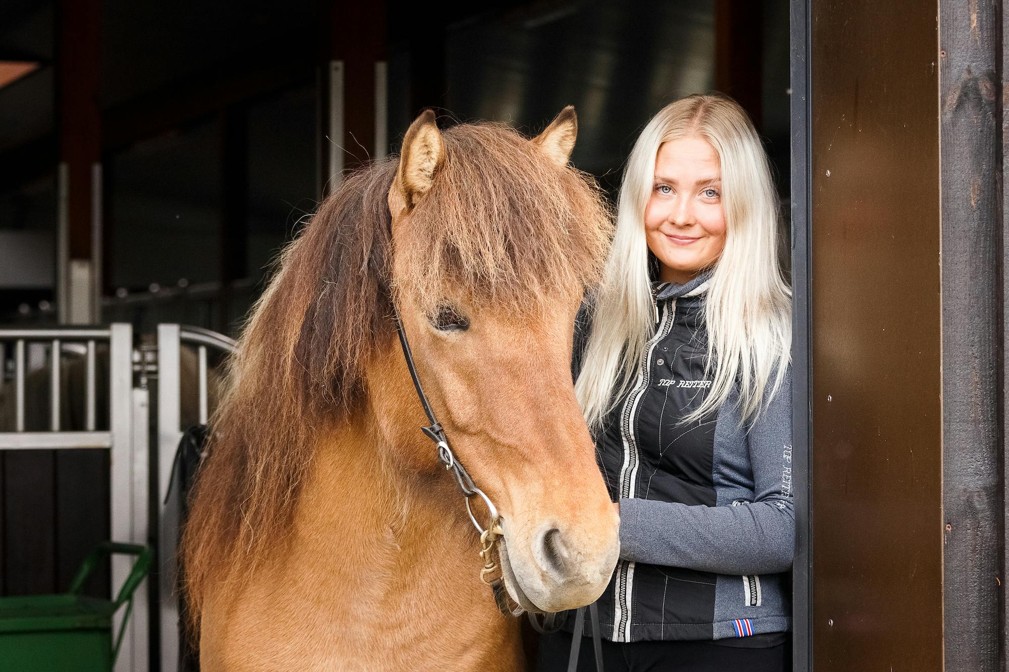 A woman standing beside an Icelandic horse in a stable doorway.