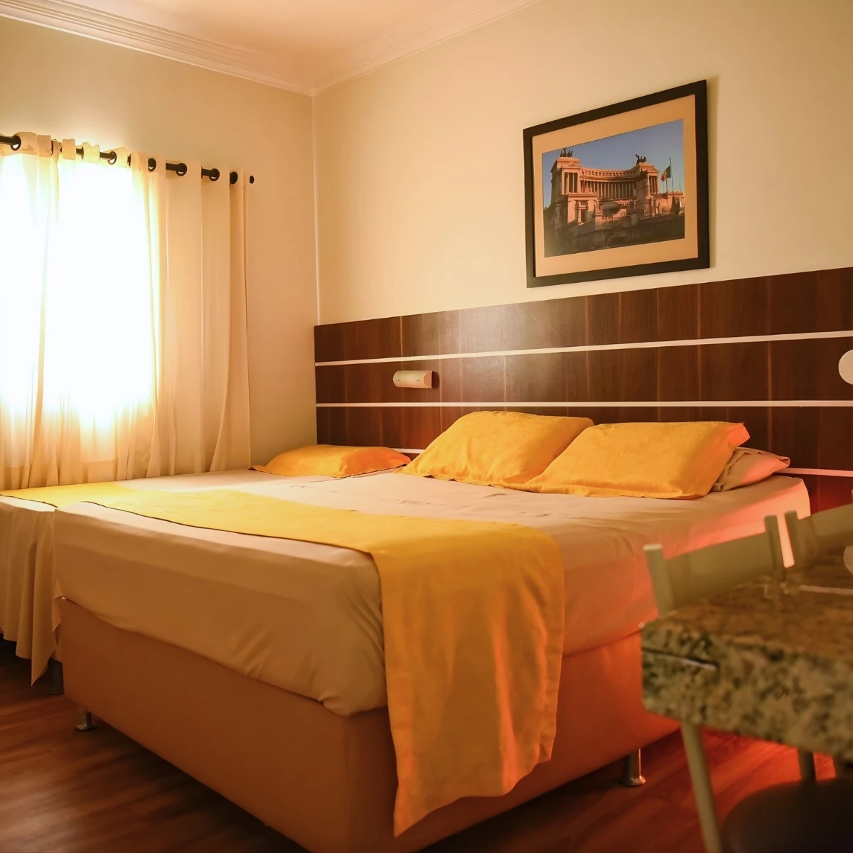 Quarto de hotel com cama de casal com lençóis e travesseiros em tons amarelados, cortinas claras na janela e quadro de prédio histórico na parede.