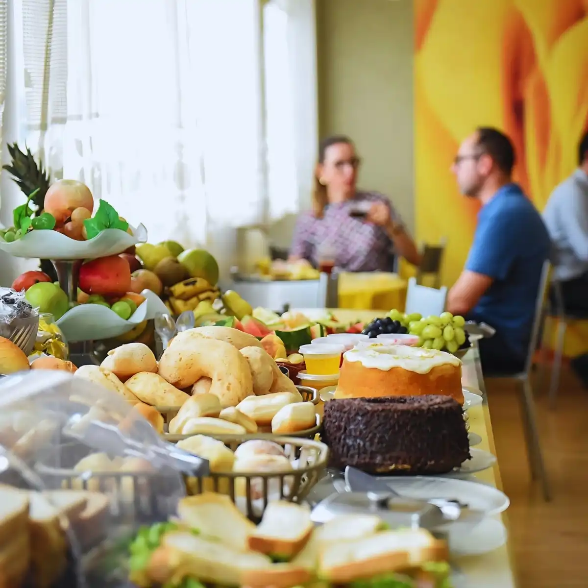 Mesa de café da manhã com pães, frutas, bolos e sucos, enquanto pessoas conversam ao fundo em ambiente iluminado, ideal para começar bem o dia.