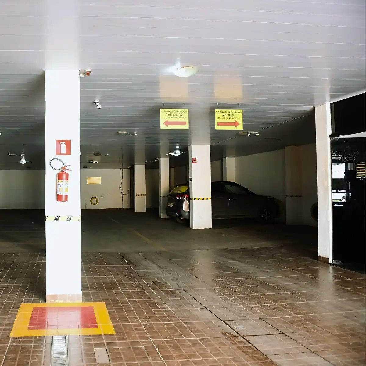 Vaga de estacionamento em garagem coberta com duas vagas destinadas a veículos pequenos, sinalizadas por placas amarelas com setas indicando a direção. Áreas de segurança marcadas no chão e extintor de incêndio disponível na parede. Ambiente limpo e organizado para estacionamento de veículos.