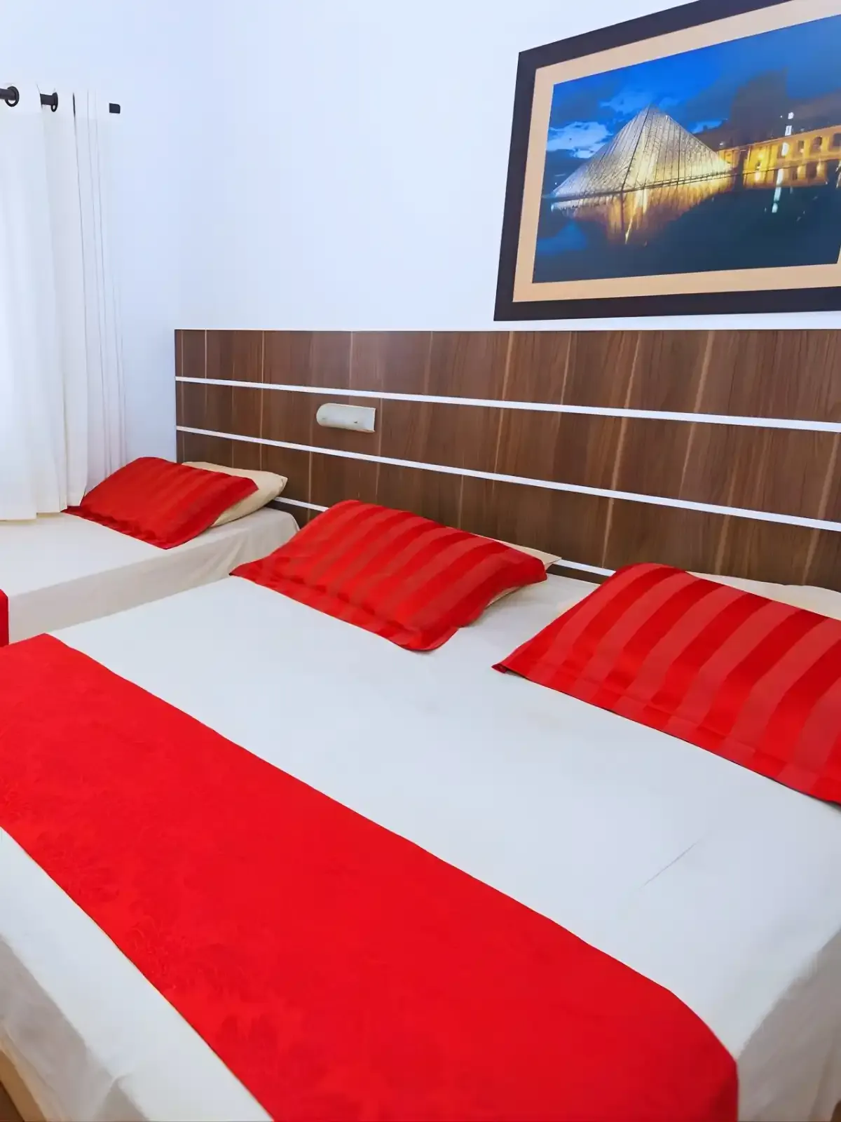 Quarto de hotel com duas camas de casal com lençóis brancos e detalhes em vermelho, cabeceira de madeira moderna e uma pintura de monumento famoso na parede.