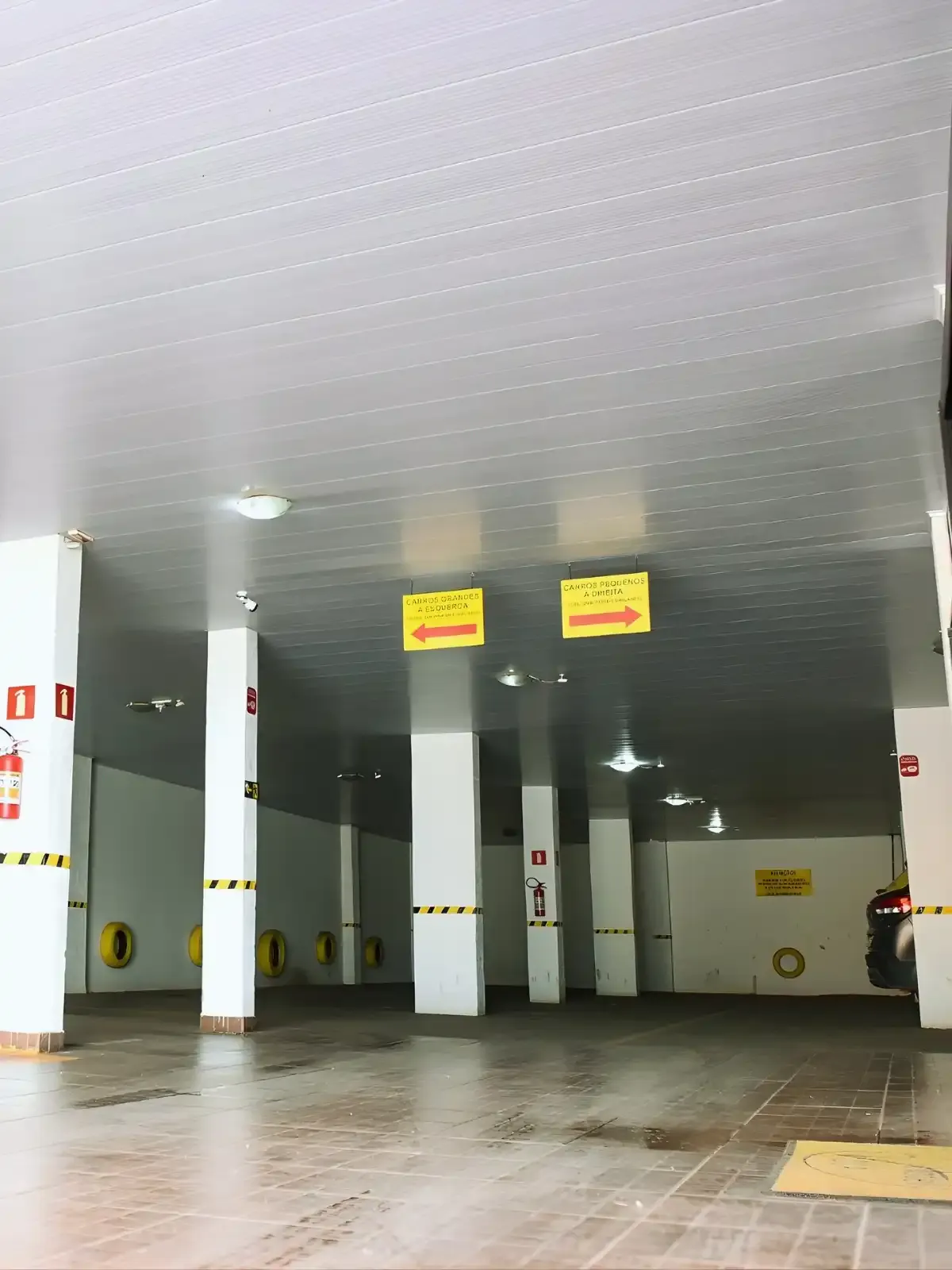 Estacionamento coberto com colunas brancas e sinalizações em amarelo e vermelho, incluindo avisos de direções e segurança, iluminação moderna.