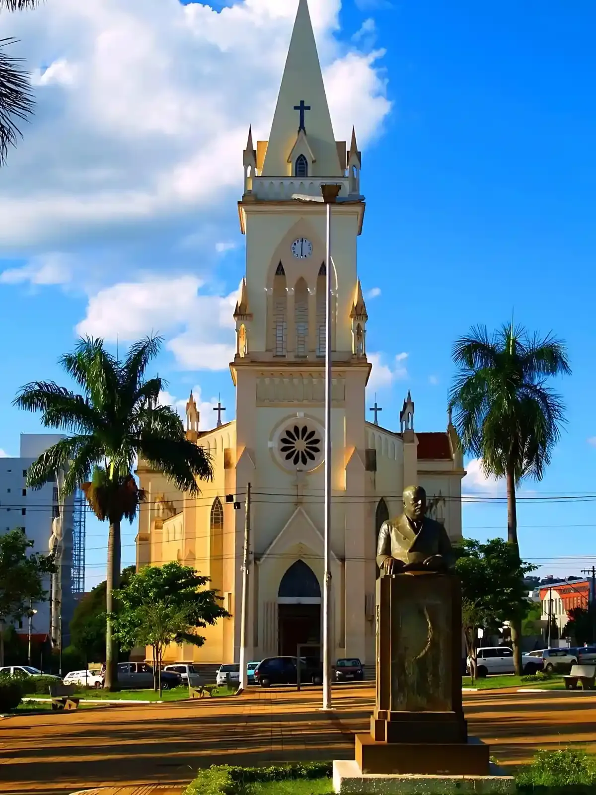 Imagem de uma igreja de estilo gótico com torre alta, cruz no topo, árvores palmadas na frente e uma estátua de uma pessoa lendo um livro, sob céu azul com nuvens brancas.