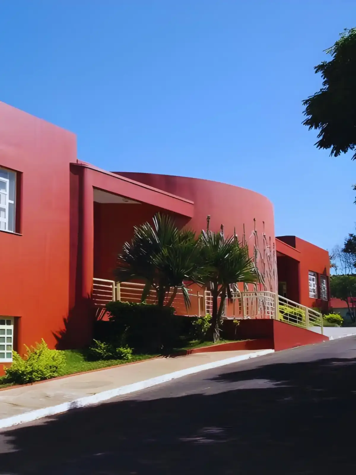 Fotografia de uma casa moderna de cor vermelha, com janelas brancas, árvores ao redor e céu azul claro ao fundo, em um bairro tranquilo.