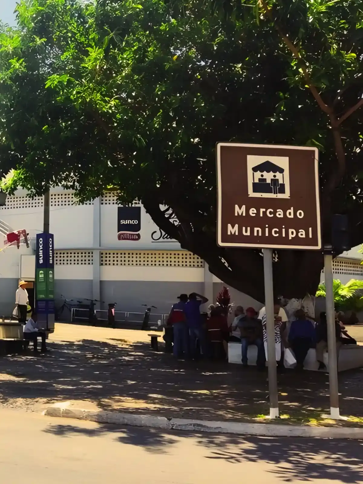 Imagem de um mercado municipal ao ar livre com várias pessoas sentadas e uma placa indicativa na frente, cercada por árvores verdes.