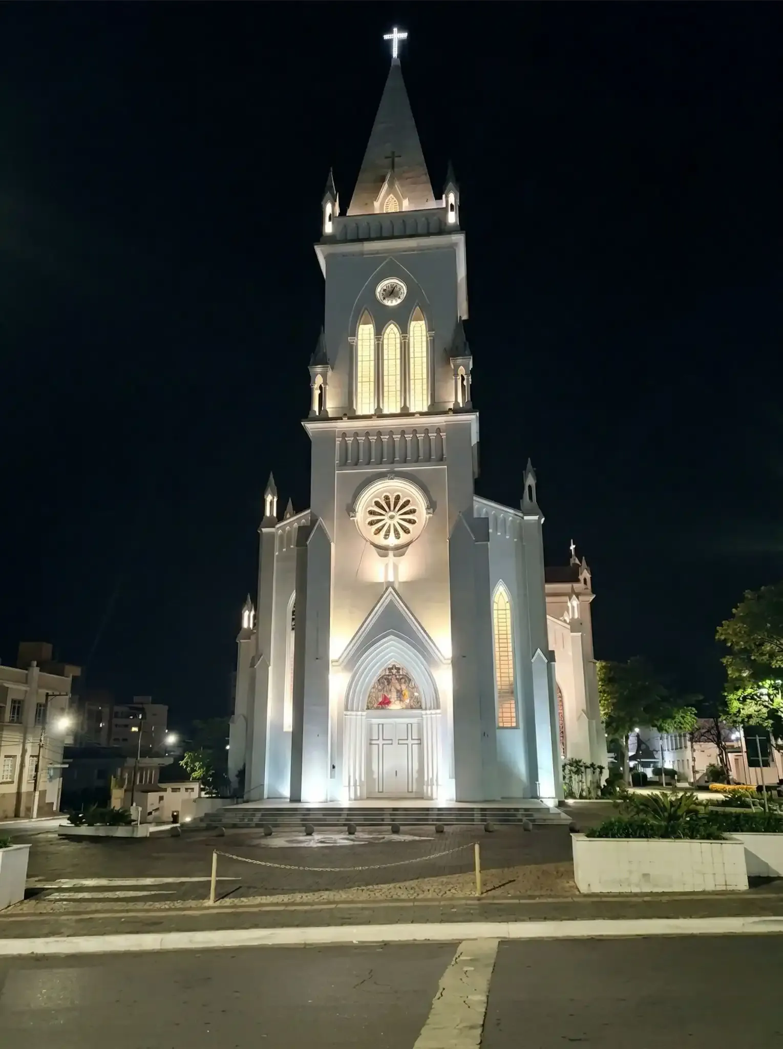 Igreja iluminada à noite, com detalhes arquitetônicos góticos e uma cruz no topo, localizada em uma praça bem iluminada.