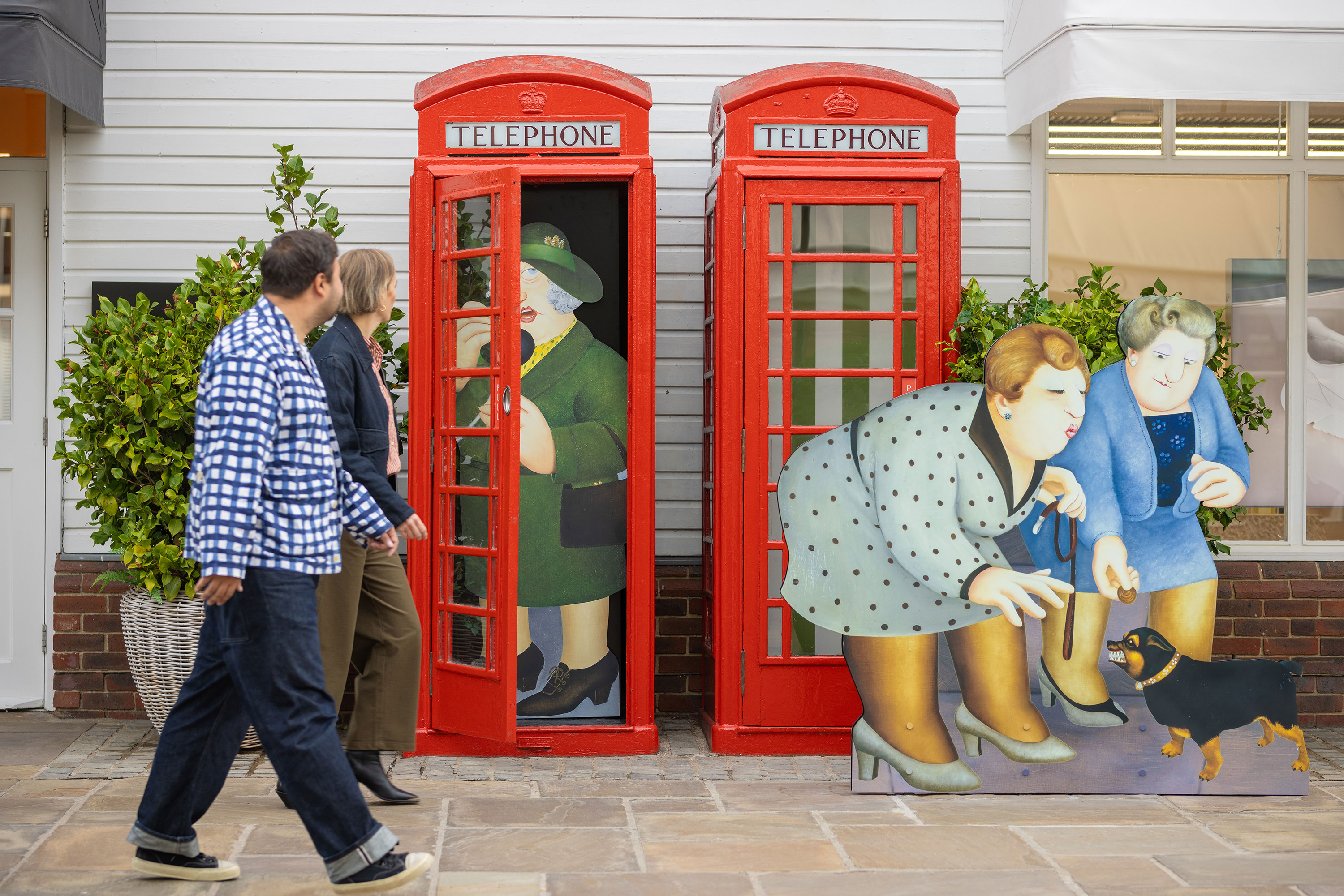 Two people walk past two classic red British phone boxes, at Bicester Village. Inside and in front, there are large cartoon-style cut-outs of people: one inside making a call, another bending to pet a small black dog, and a woman standing nearby. Green shrubs sit behind the boxes.