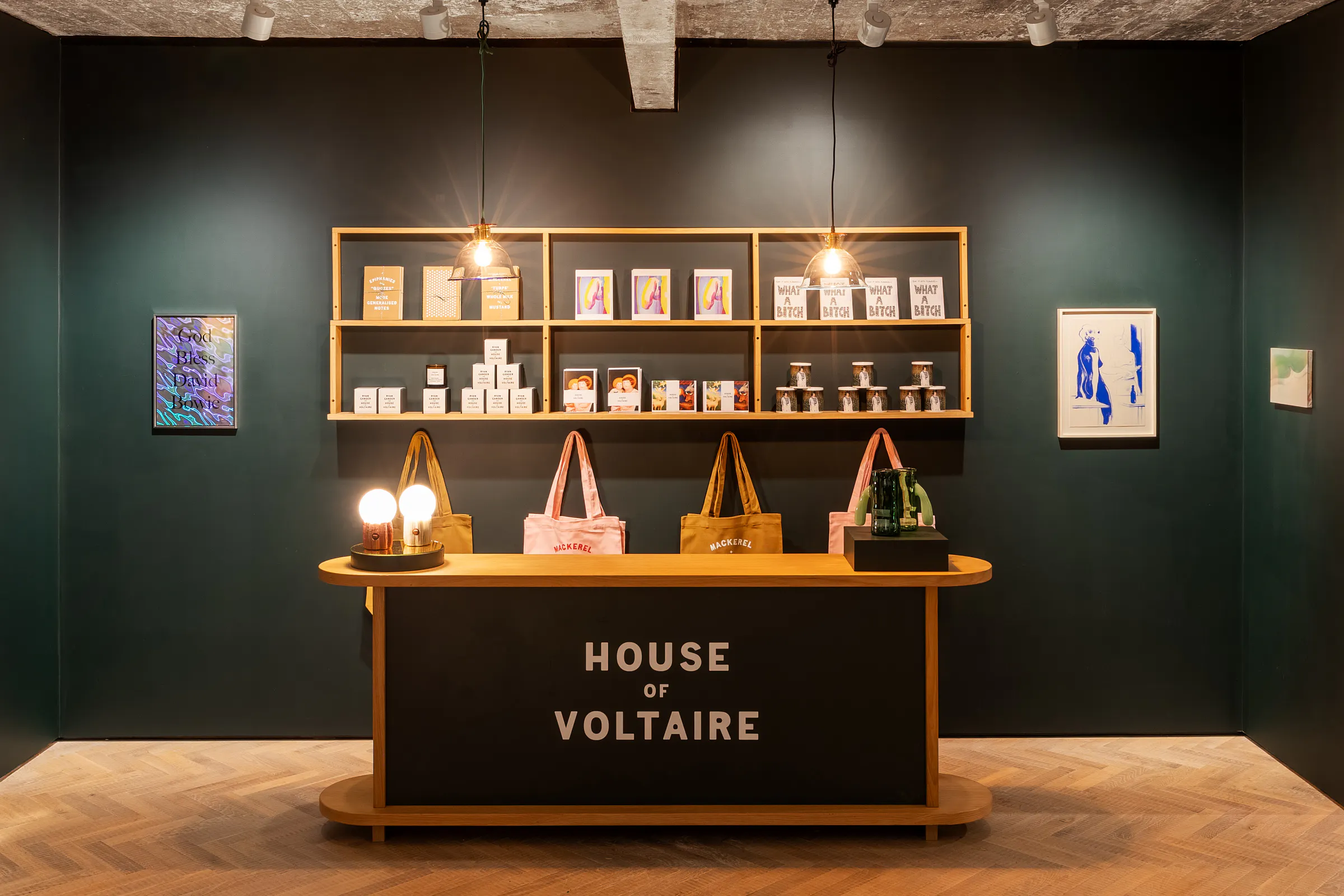 A stylish, modern pop-up shop display featuring a dark green wall with framed art and shelves holding books and items. A wooden counter with the text "House of Voltaire" is in front, with tote bags and lights adding a warm ambiance.