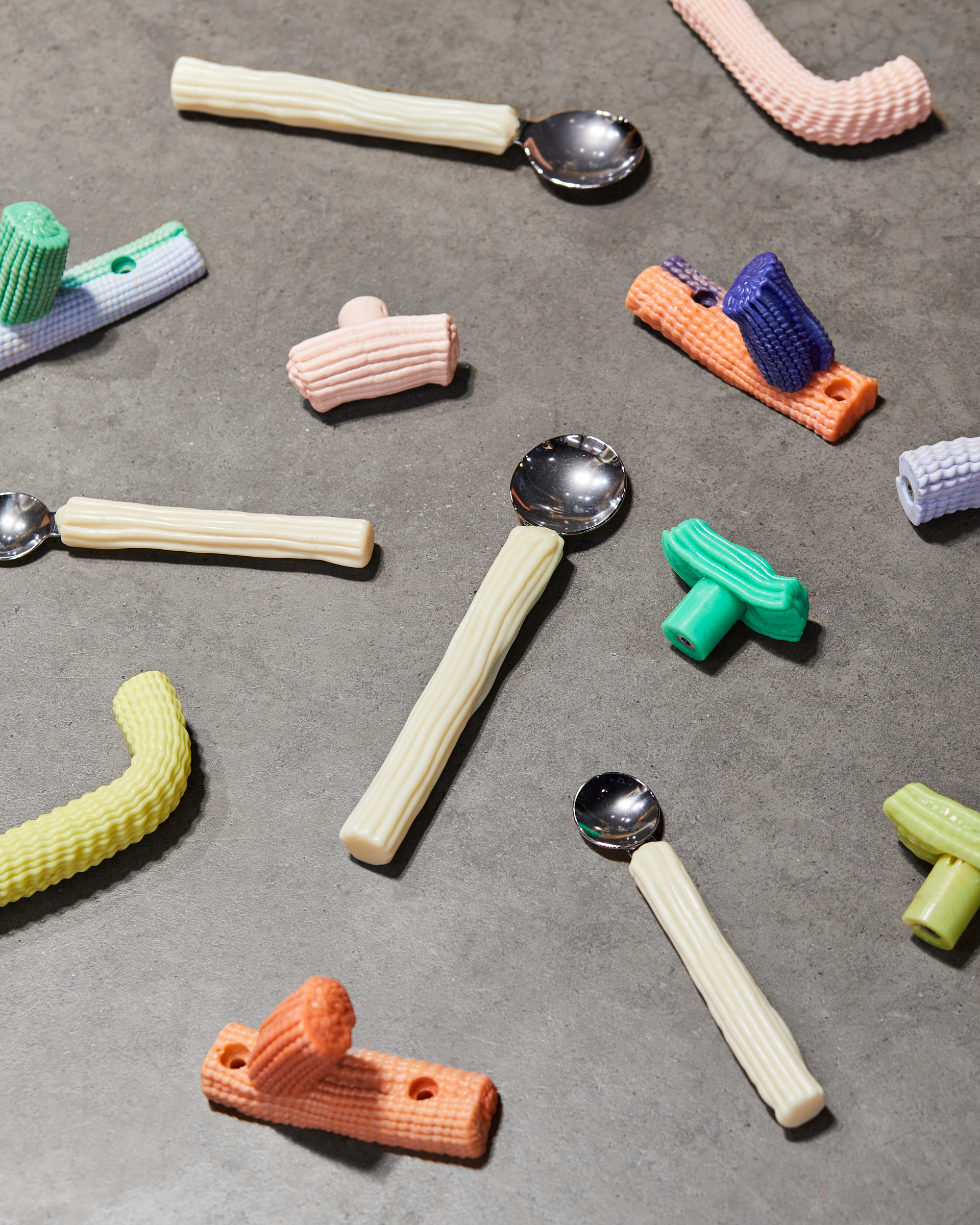 Assorted colourful spoons and plastic-coated handles and clothes hooks arranged on a grey surface. The spoons, handles and clothes hooks are ribbed in different colourful shades, creating a playful and artistic composition.