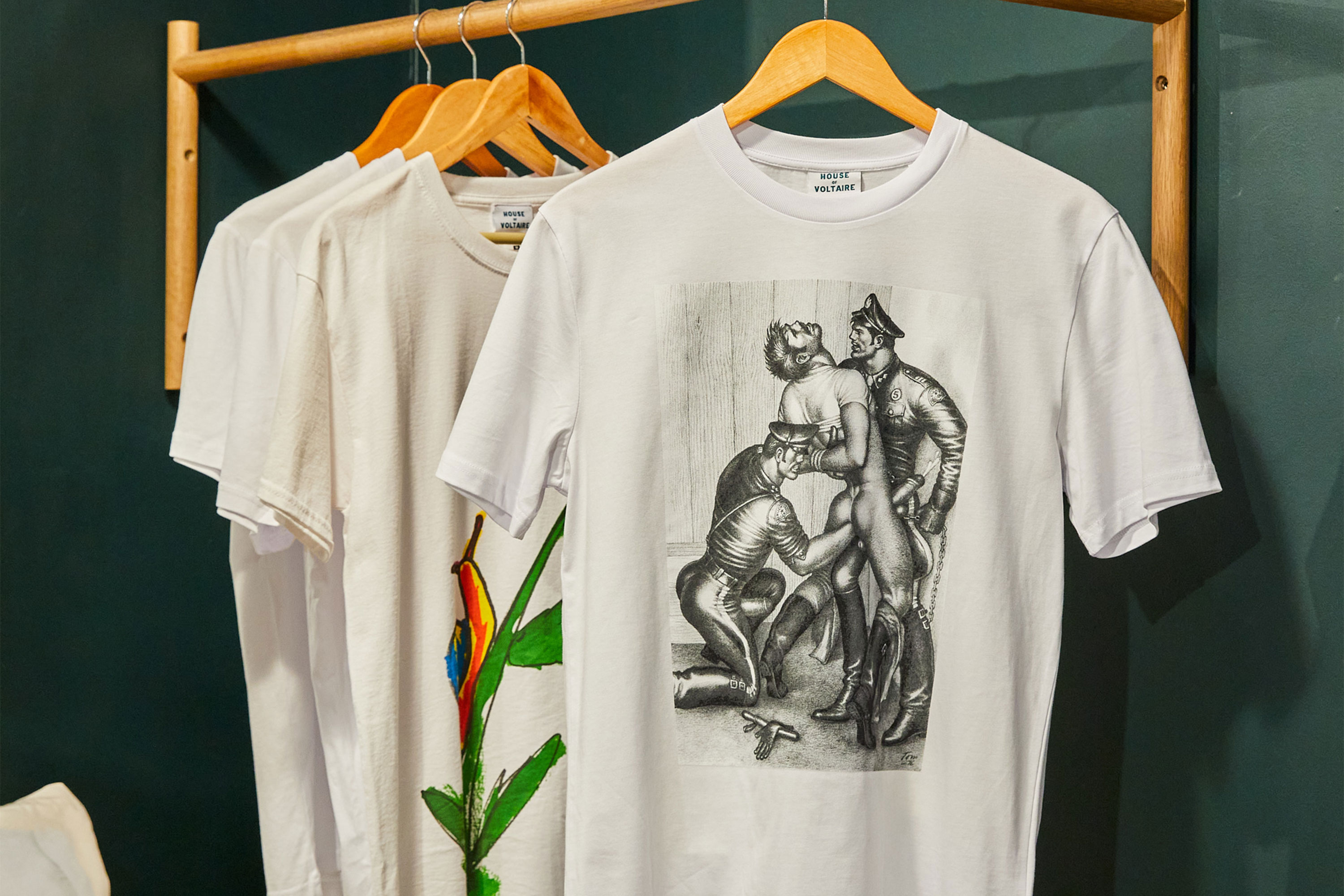 Three white t-shirts hang on wooden hangers; one features a colourful snail and plant design, another has a greyscale illustration by Tom of Finland, and the third is mostly obscured.