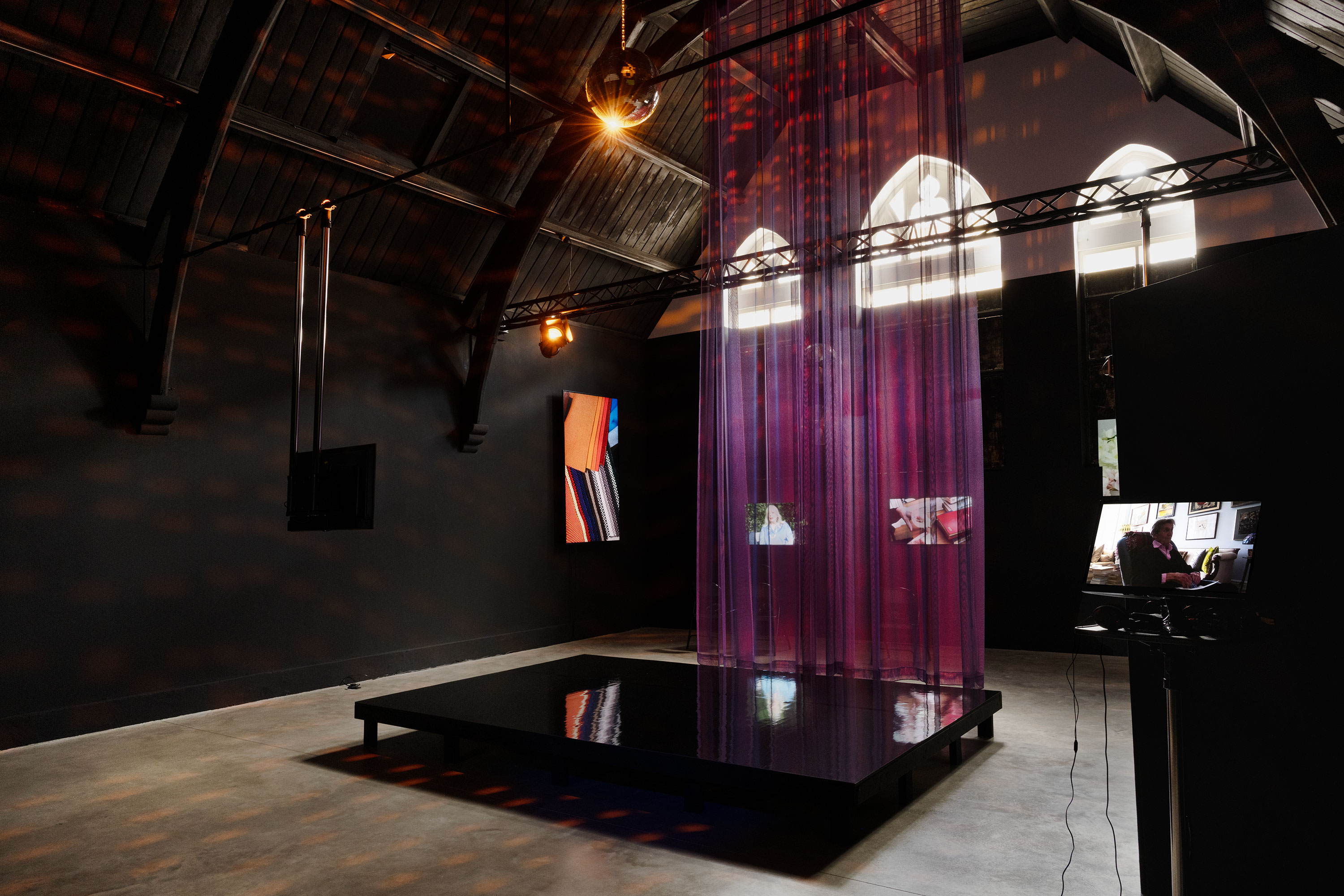 A dark, high-ceilinged gallery with arched windows features a glossy black platform topped with sheer purple curtains. Spotlights cast warm and coloured reflections. Mounted screens display abstract and close-up images along the surrounding black walls.