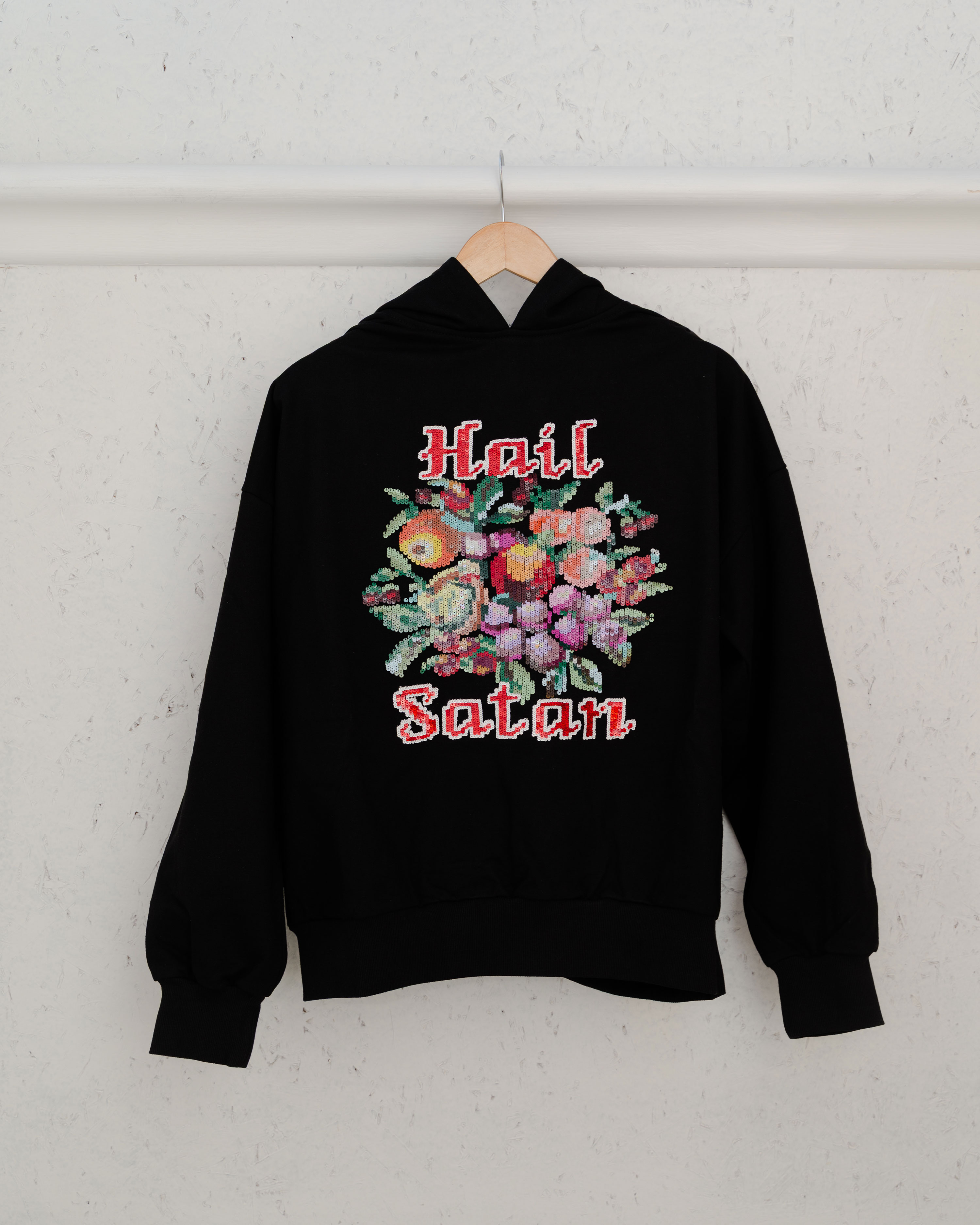 A black hoodie hangs by designer Ashish Gupta on a white wall. Its back features a colourful embroidered floral design with the words "Hail Satan." in large, white, and red outlined cross-stitch style lettering above and below the flowers.