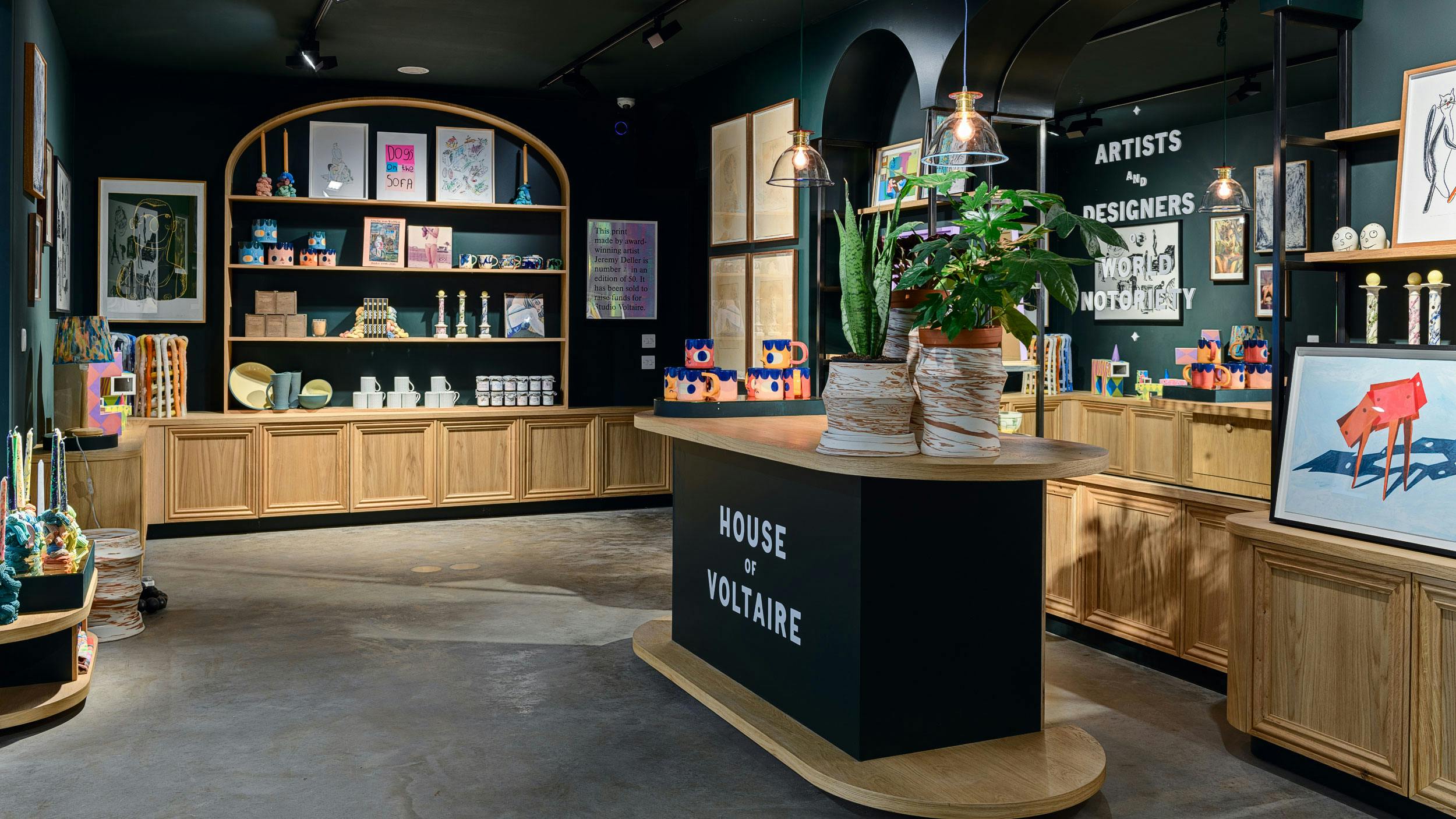 An interior photograph of the House of Voltaire store. The walls are painted a dark green, and in built shelving Ash wood shelving line the walls. The shelving is filled with uniquer artworks and homeware objects.