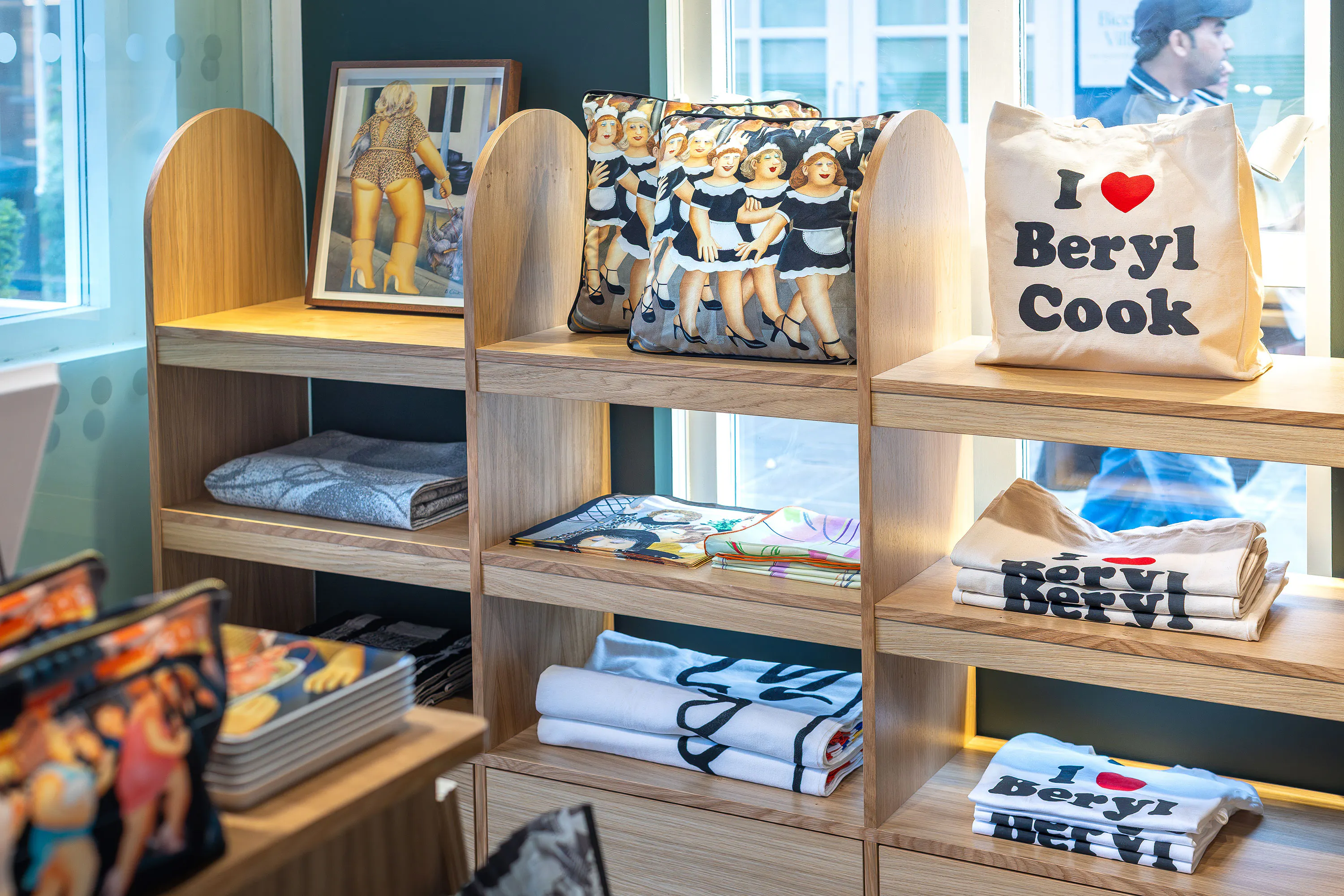 Wooden shelves display Beryl Cook-themed merchandise including tote bags, cushions with cartoon women, folded T-shirts, books, and a framed print featuring dancing women, all brightly lit by large windows in a modern shop interior.