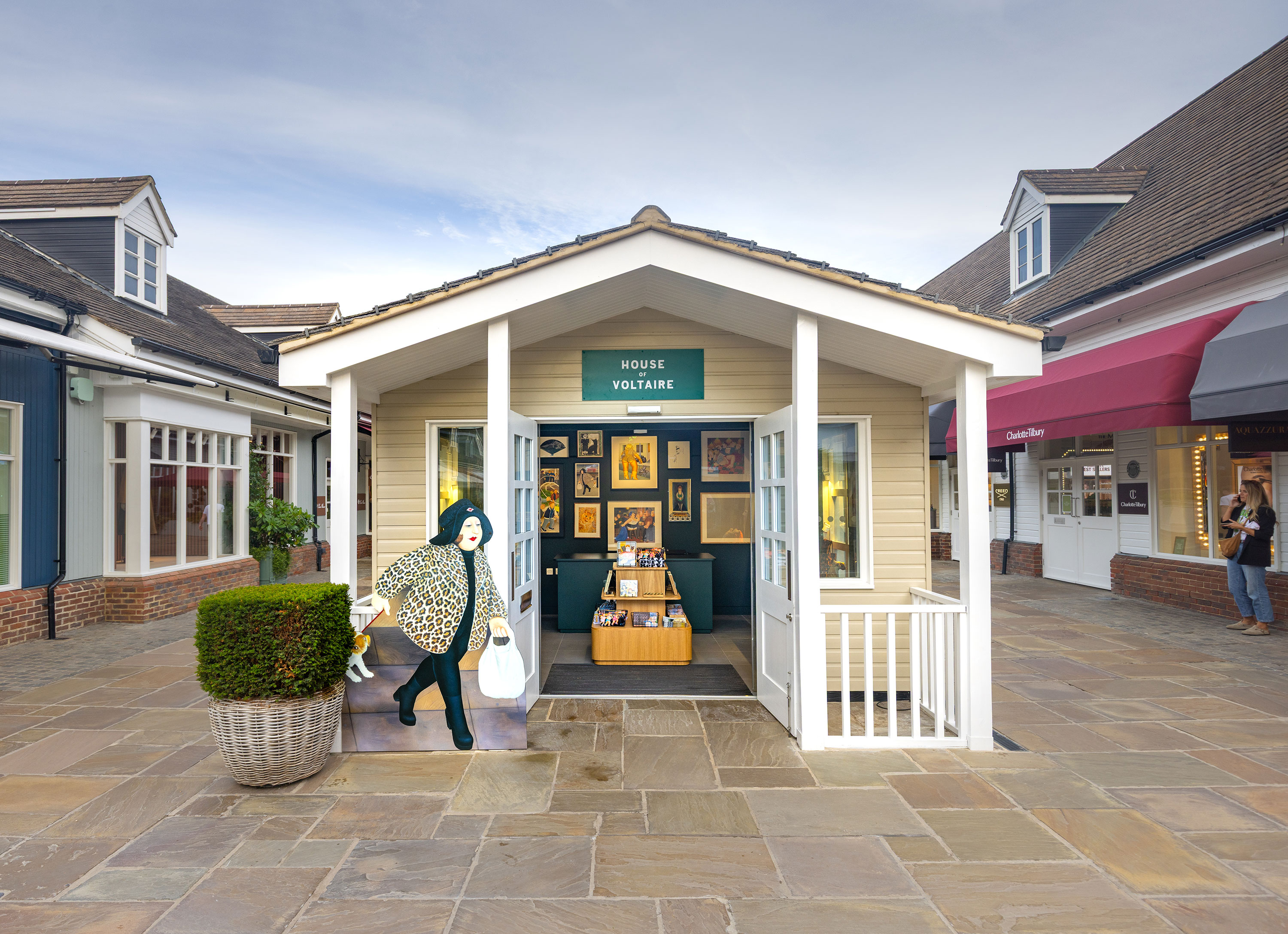 Small beige house-style shop labelled “House of Voltaire.” Artworks fill the back wall. A life-size Beryl Cook character with a leopard coat stands beside a potted shrub. Shops line the outdoor plaza, and a person walks in the background.