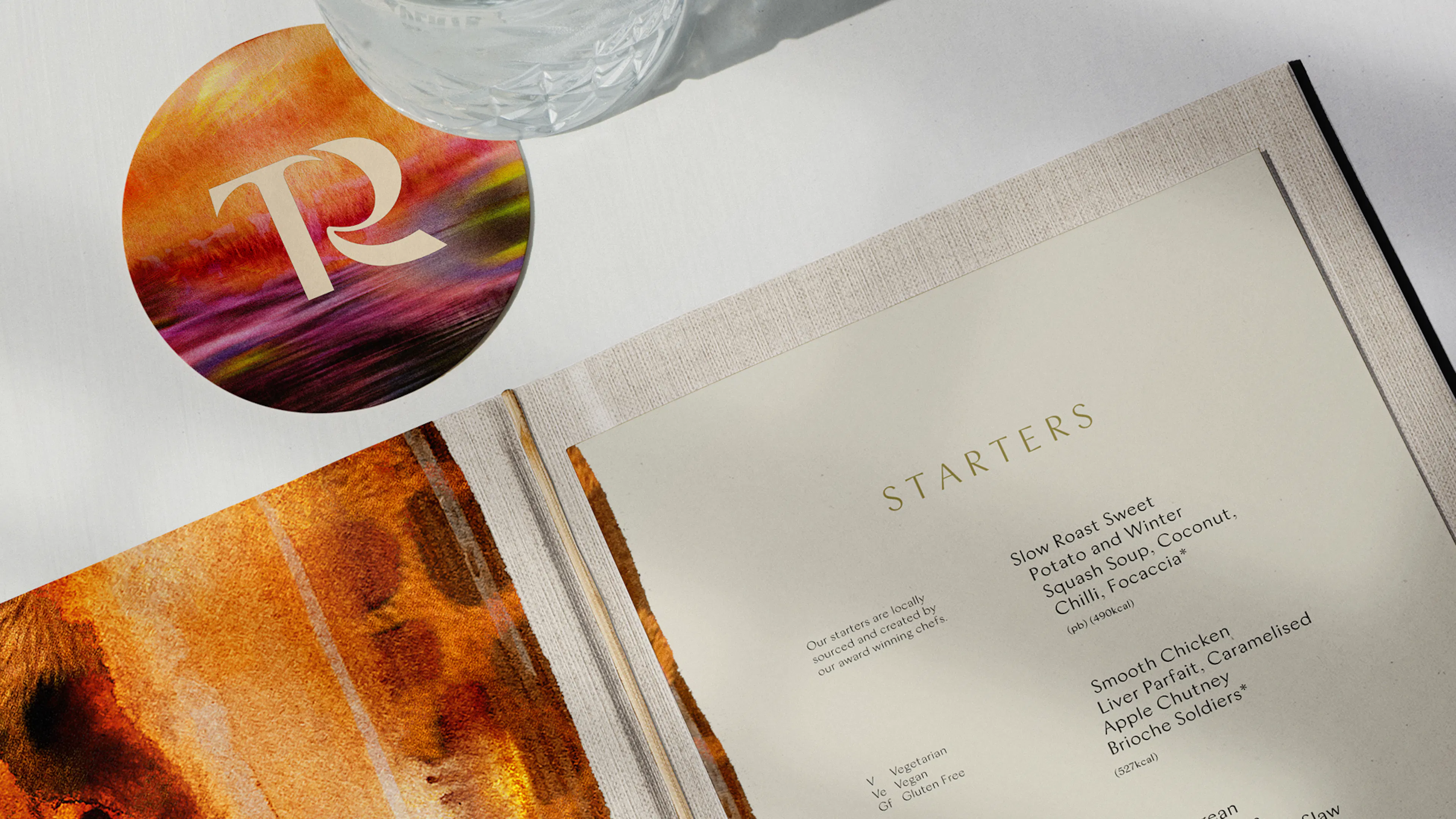 Open restaurant menu showing a Starters page, with watercolour endpapers and a round TR coaster under a glass
