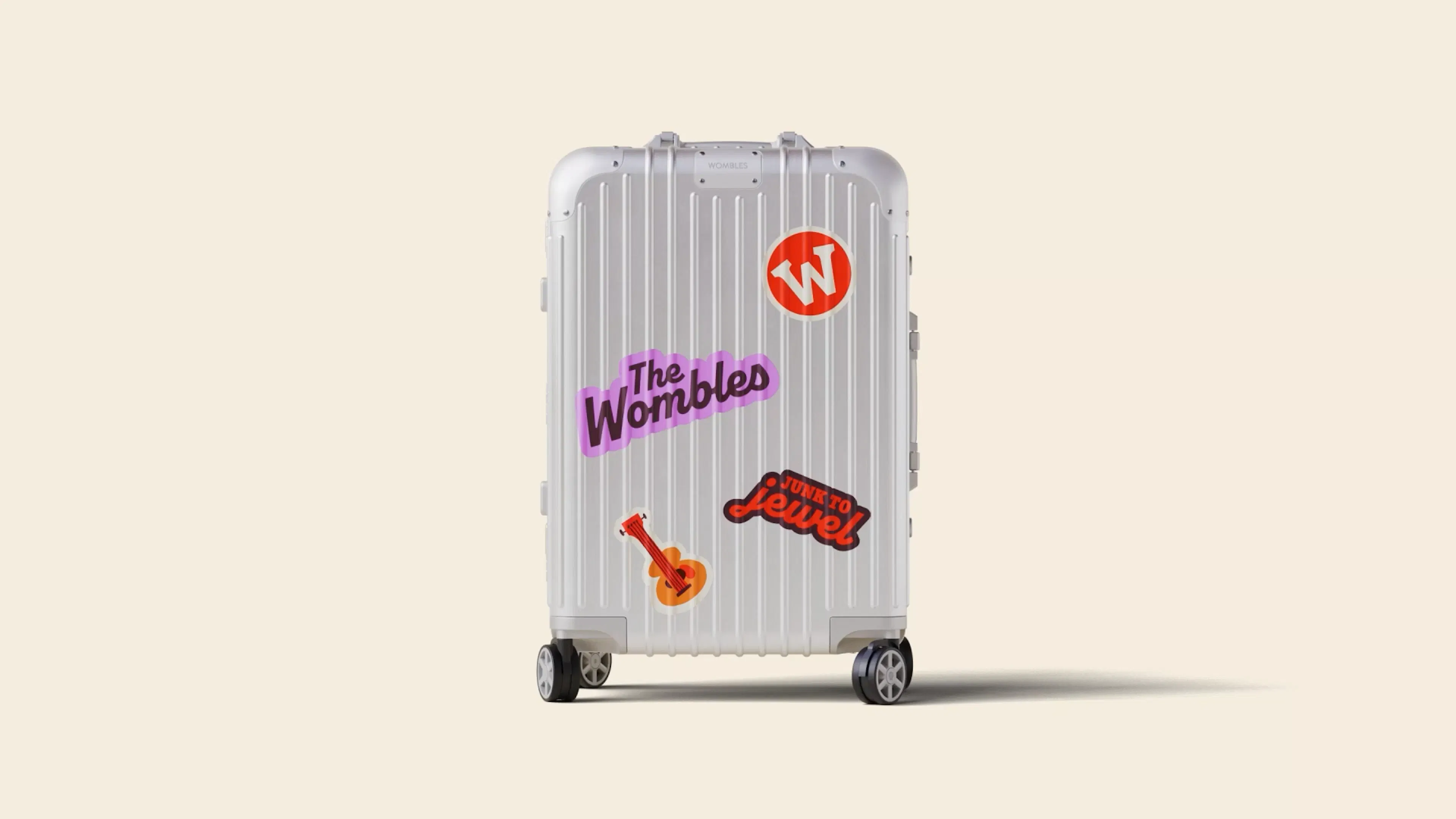 A silver hard-shell suitcase on a cream background, decorated with The Wombles stickers including the red W logo, a purple wordmark and a guitar