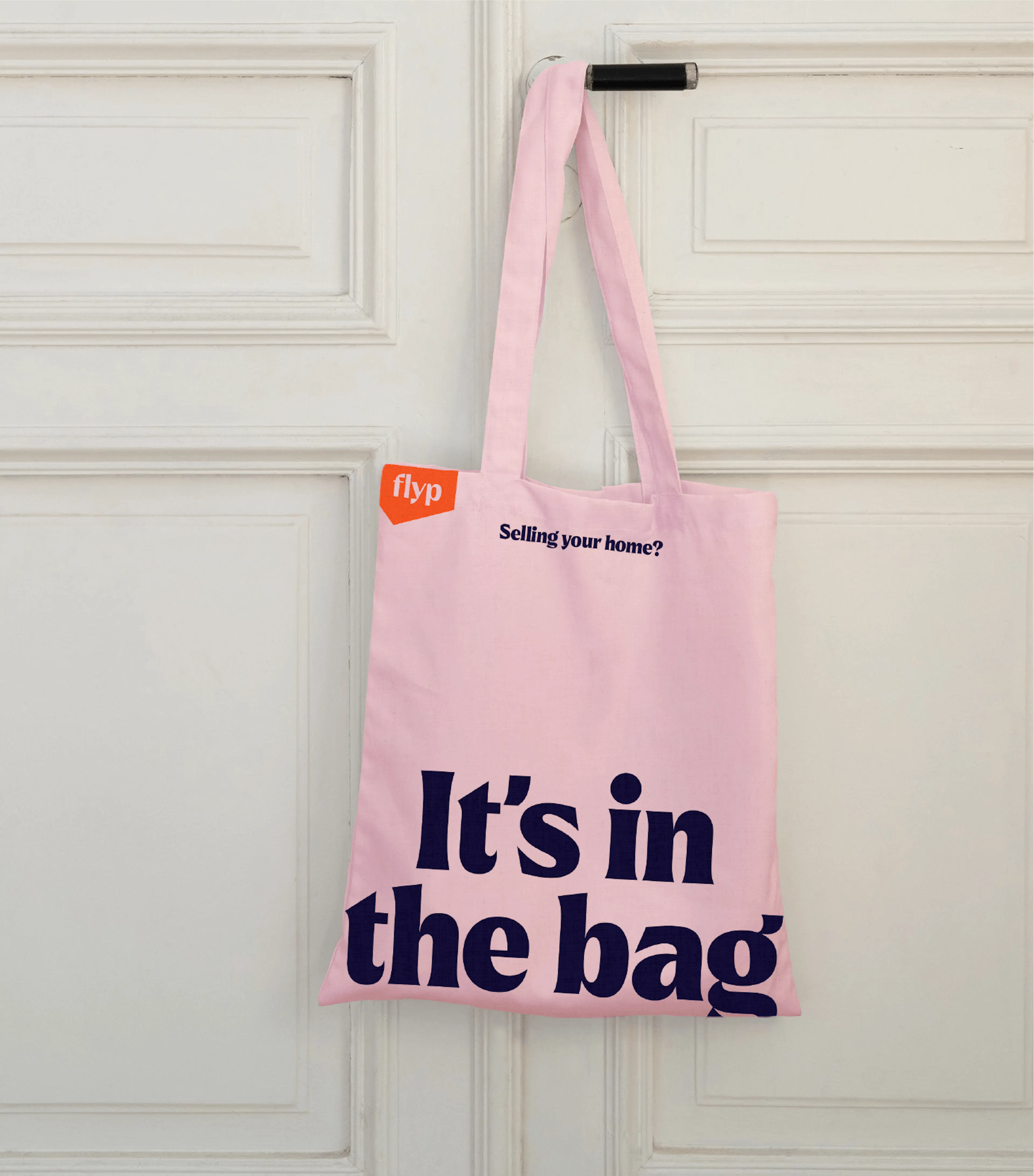 Pink flyp tote bag reading 'It's in the bag' hanging on a white door