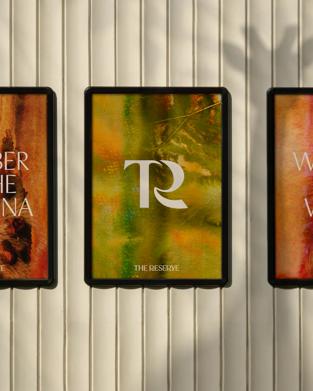 Framed poster of the TR monogram over a green and gold watercolour pattern on a panelled wall, flanked by two more posters