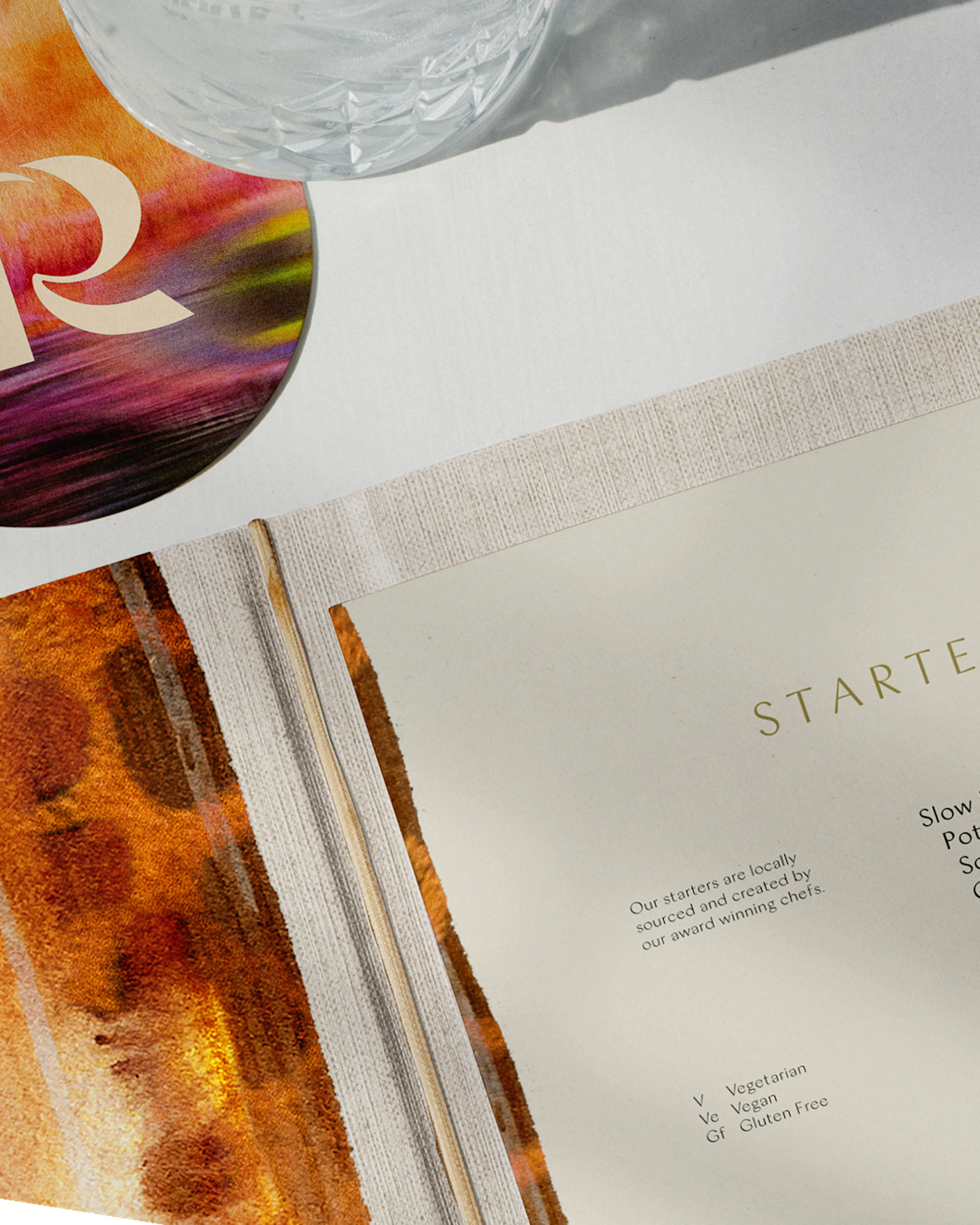 Open restaurant menu showing a Starters page, with watercolour endpapers and a round TR coaster under a glass